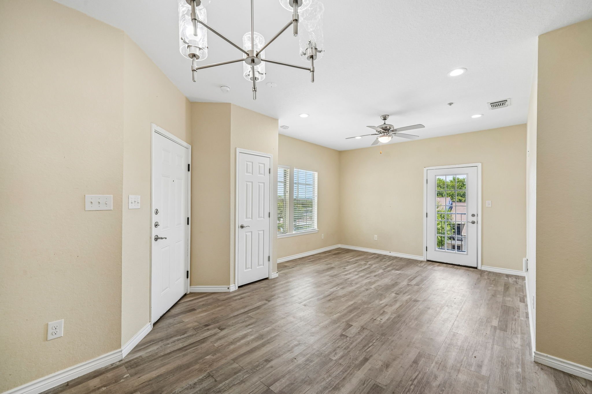 An empty living room with beige walls, wood flooring, a white ceiling fan, and a modern chandelier. There are several windows and a door leading outside, with white trim around the windows and doors.