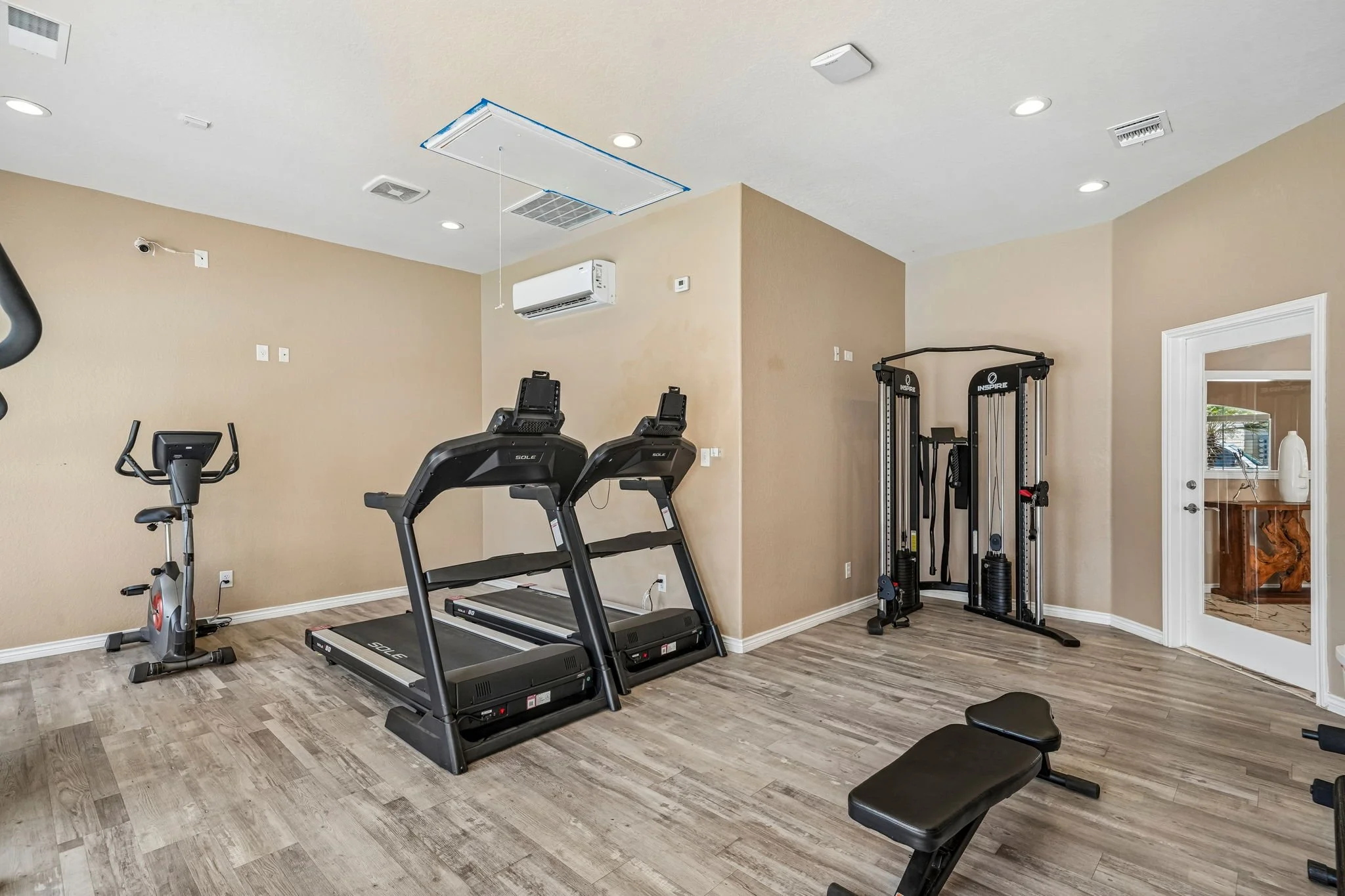 24-hour gym in the clubhouse at La Marquesa Apartments with elliptical, treadmill, weight machine, bench, and TV, with beige walls, wood floor, and ceiling lighting.