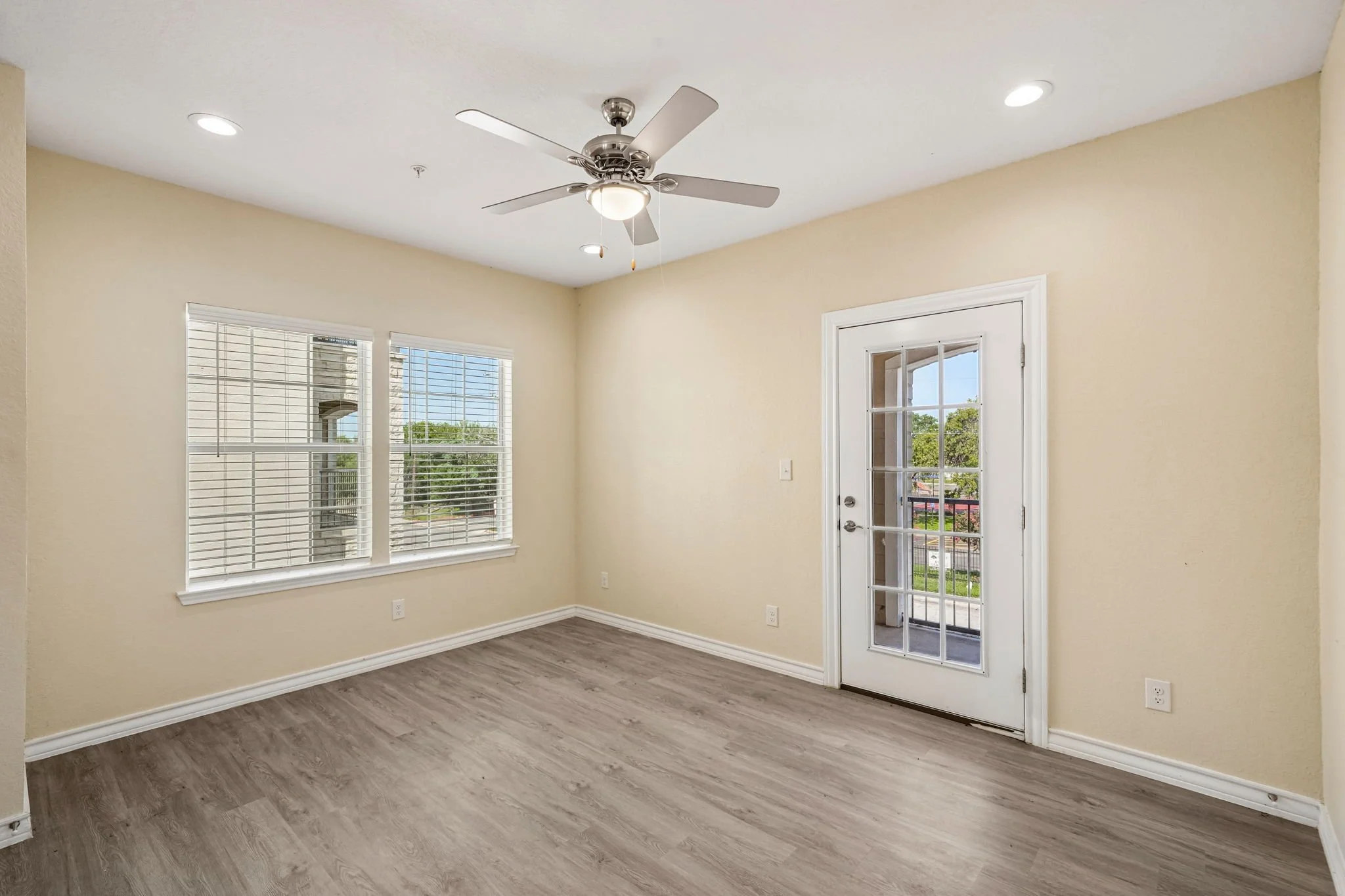 Empty living room with beige walls, wood laminate flooring, three windows with white blinds, a glass-paneled door leading outside, ceiling fan with light, and recessed ceiling lights.