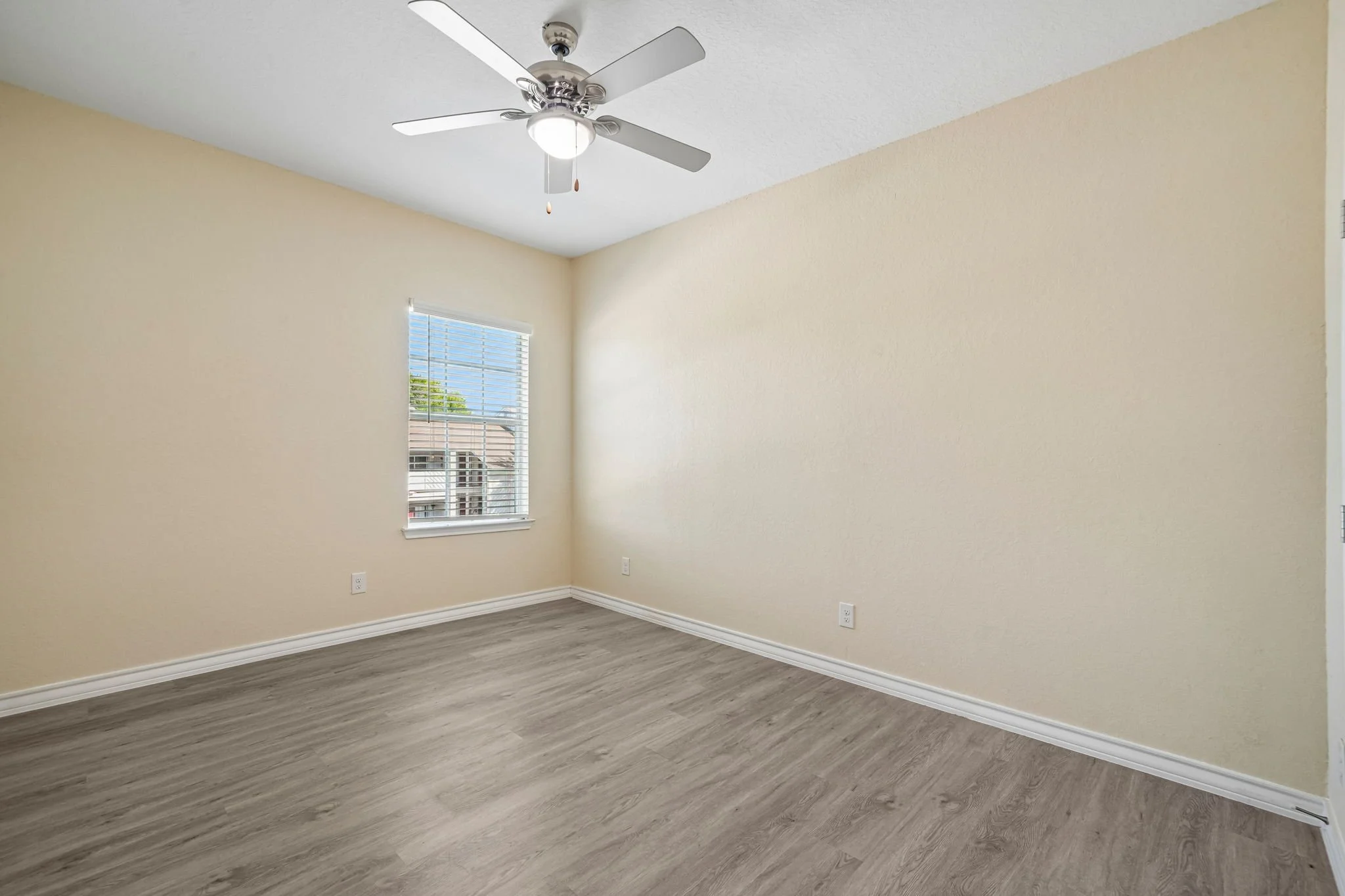 Empty bedroom with beige walls, a window with blinds, a ceiling fan with a light, and wood-look flooring.