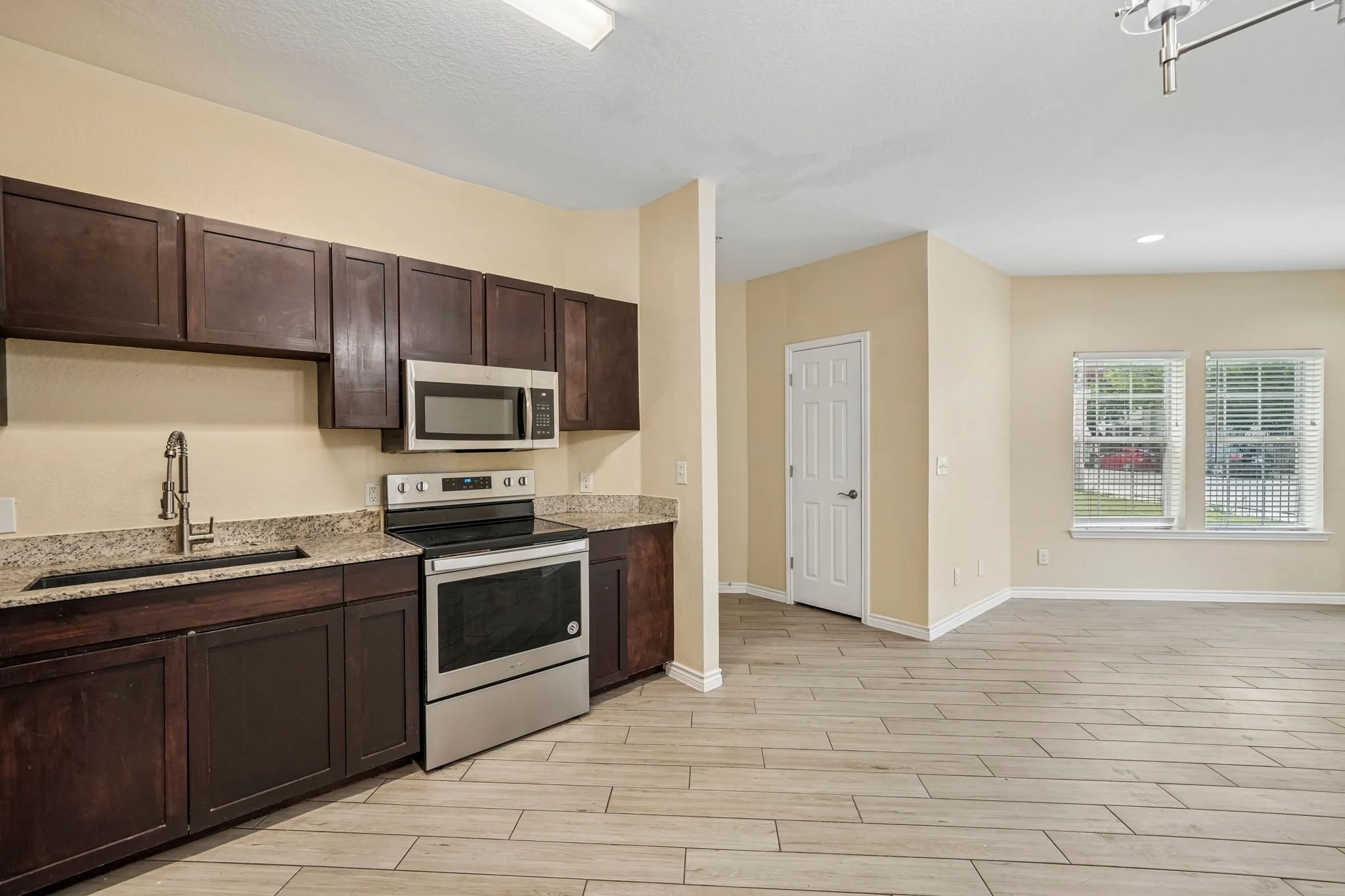 Interior view of a modern kitchen with dark wooden cabinets, a stainless steel microwave oven, an electric stove, granite countertops, and a faucet over the sink. Adjacent to the kitchen is a bright living space with light-colored walls, a white door