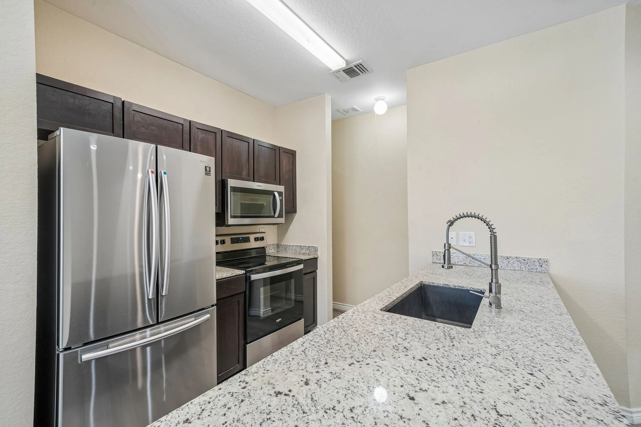 Kitchen with granite countertop, stainless steel refrigerator, microwave, oven, dark wooden cabinets, and a black sink with a silver faucet.