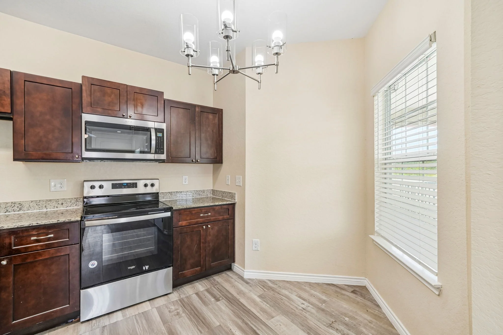 A kitchen corner with wooden cabinetry, a stainless steel microwave oven, and an electric stove with a black glass top. There is a window with white blinds on the right side, and a modern chandelier hanging from the ceiling.