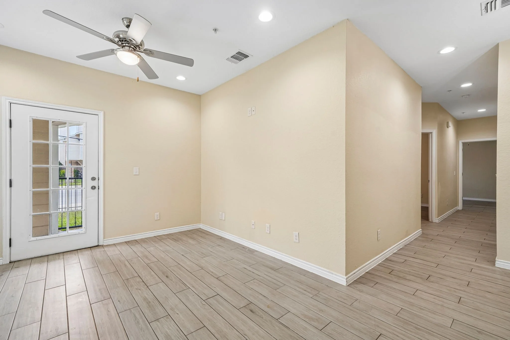 Empty living room with beige walls, wooden flooring, a door with glass panels leading outside, ceiling fan with light, and recessed lighting and a hallway leading to other rooms in the unit. 