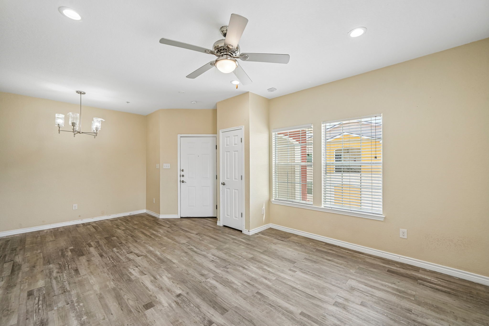 Empty living room with beige walls, wood-look flooring, white trim, three windows with blinds, a white door, and two ceiling lights including a ceiling fan with a light.