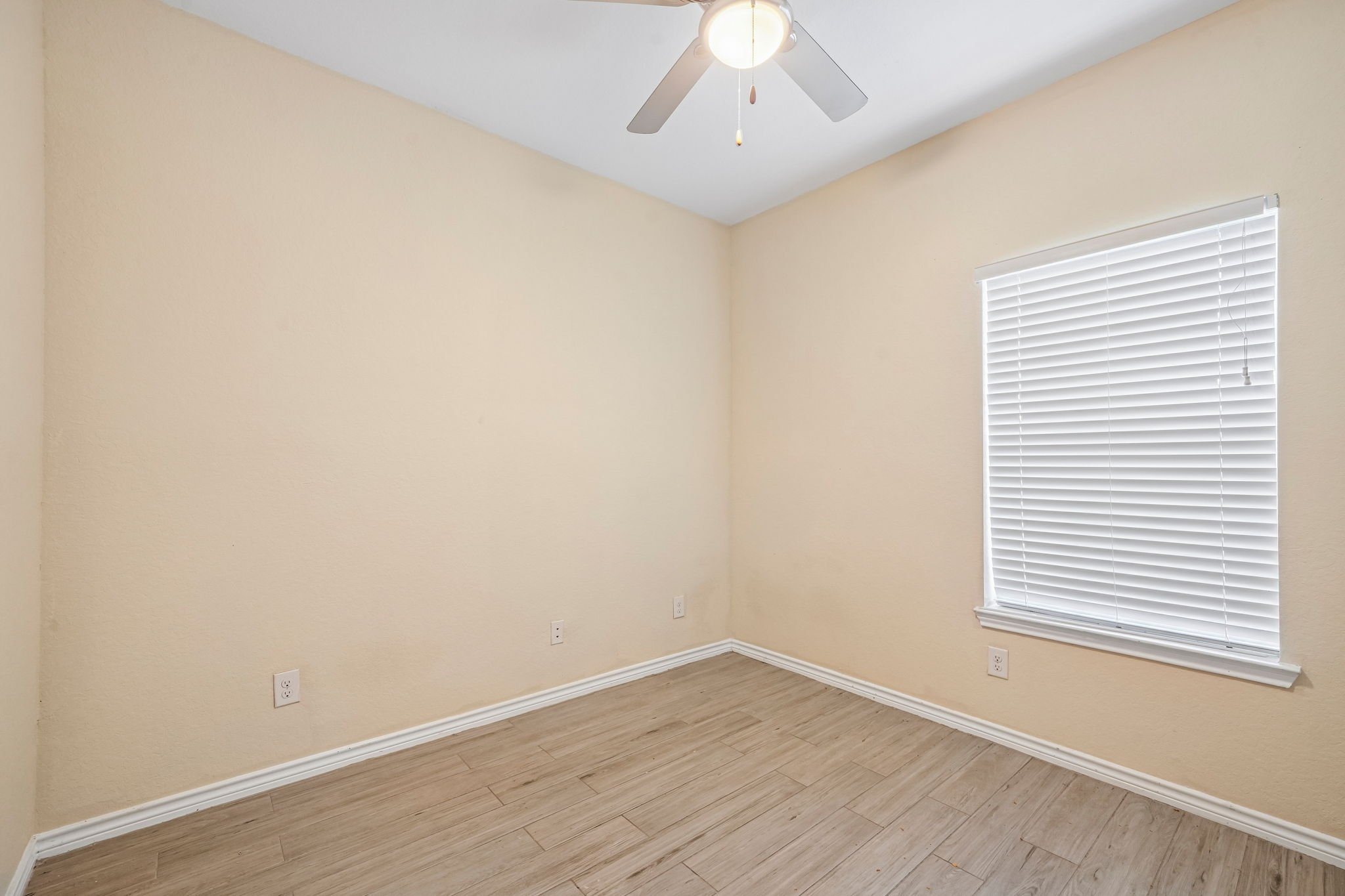 Empty bedroom with beige walls, white window blinds, ceiling fan with light, and wood-look flooring.