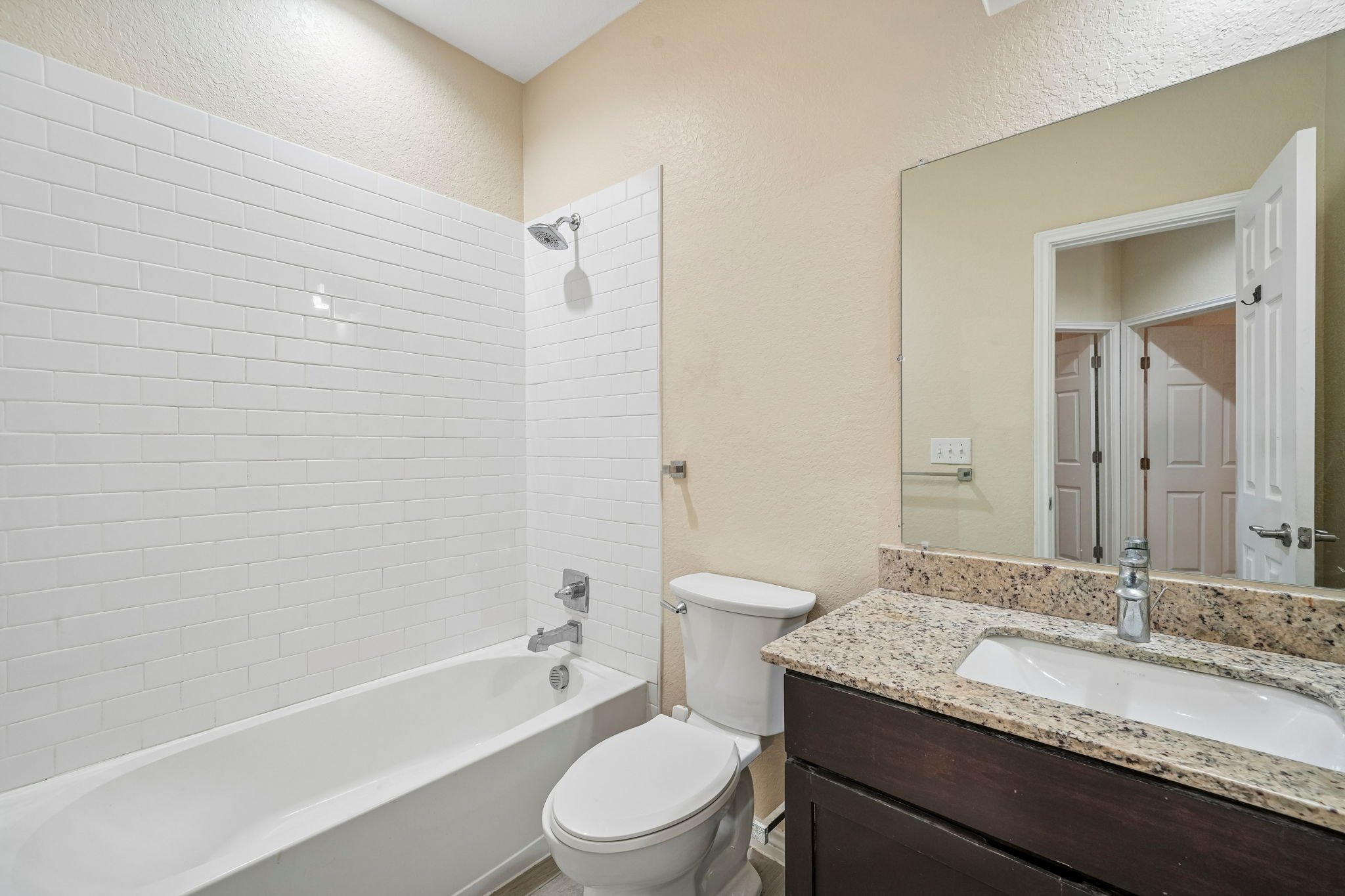 Bathroom with a bathtub and shower, white subway tiles, toilet, granite countertop with sink, mirror, and closed door.