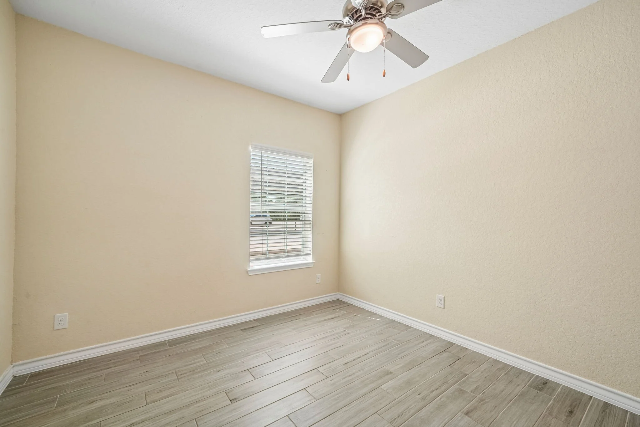 Empty bedroom with beige walls, a window with blinds, ceiling fan, and wood-looking flooring.
