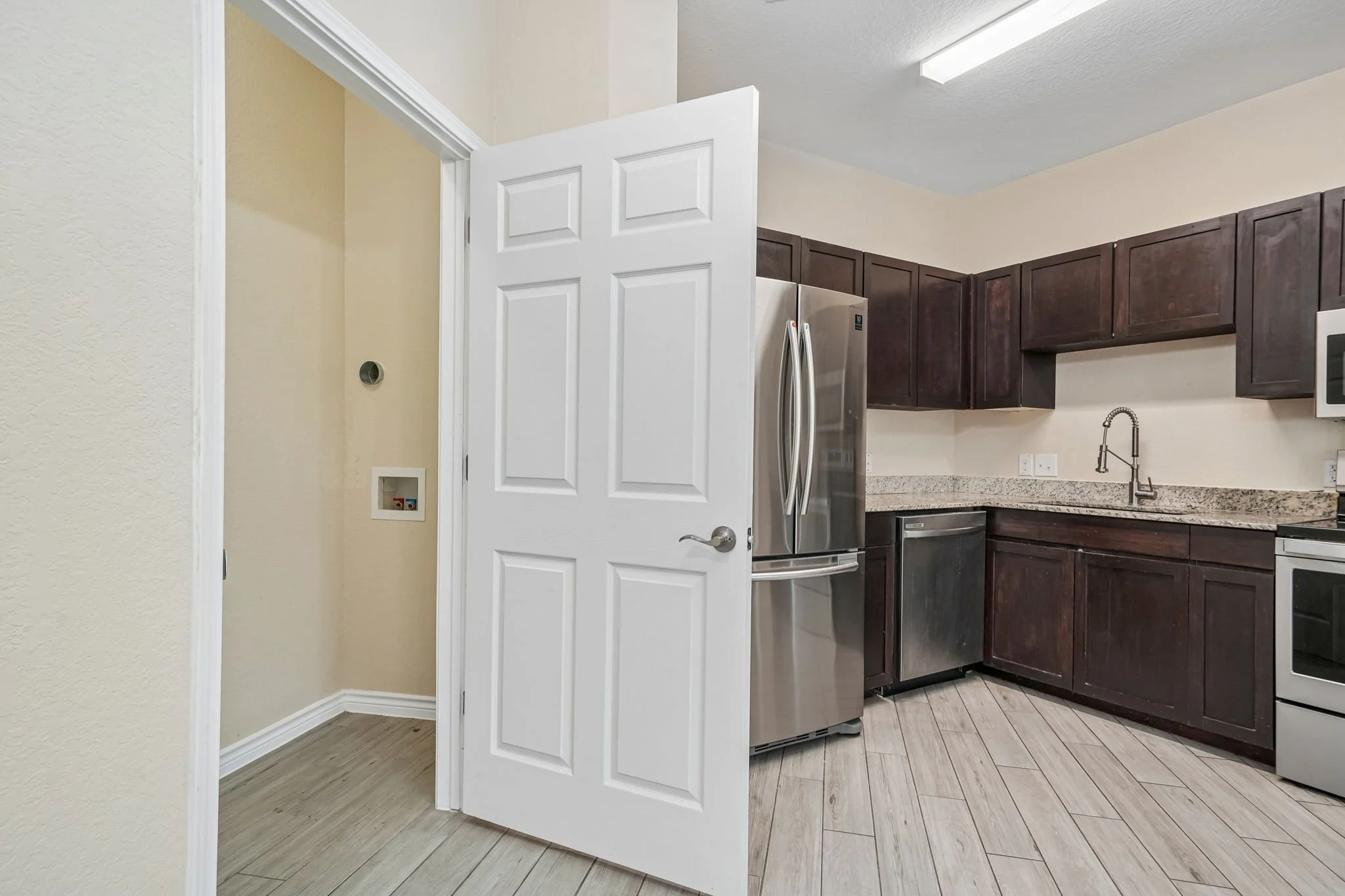 A kitchen with dark brown cabinets, a stainless steel refrigerator, white walls, a granite countertop, a stainless steel sink with a modern faucet, and a microwave. An open white door reveals a small laundry area with in-unit utilities.