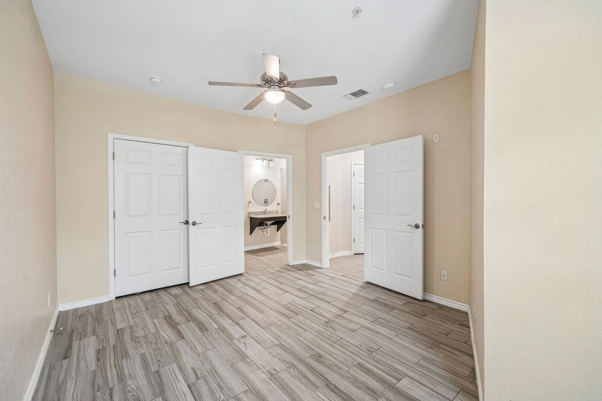 Empty primary bedroom with light-colored walls, gray wood-look tile floor, and white double doors leading to a bathroom with a sink and mirror. Ceiling fan with light.