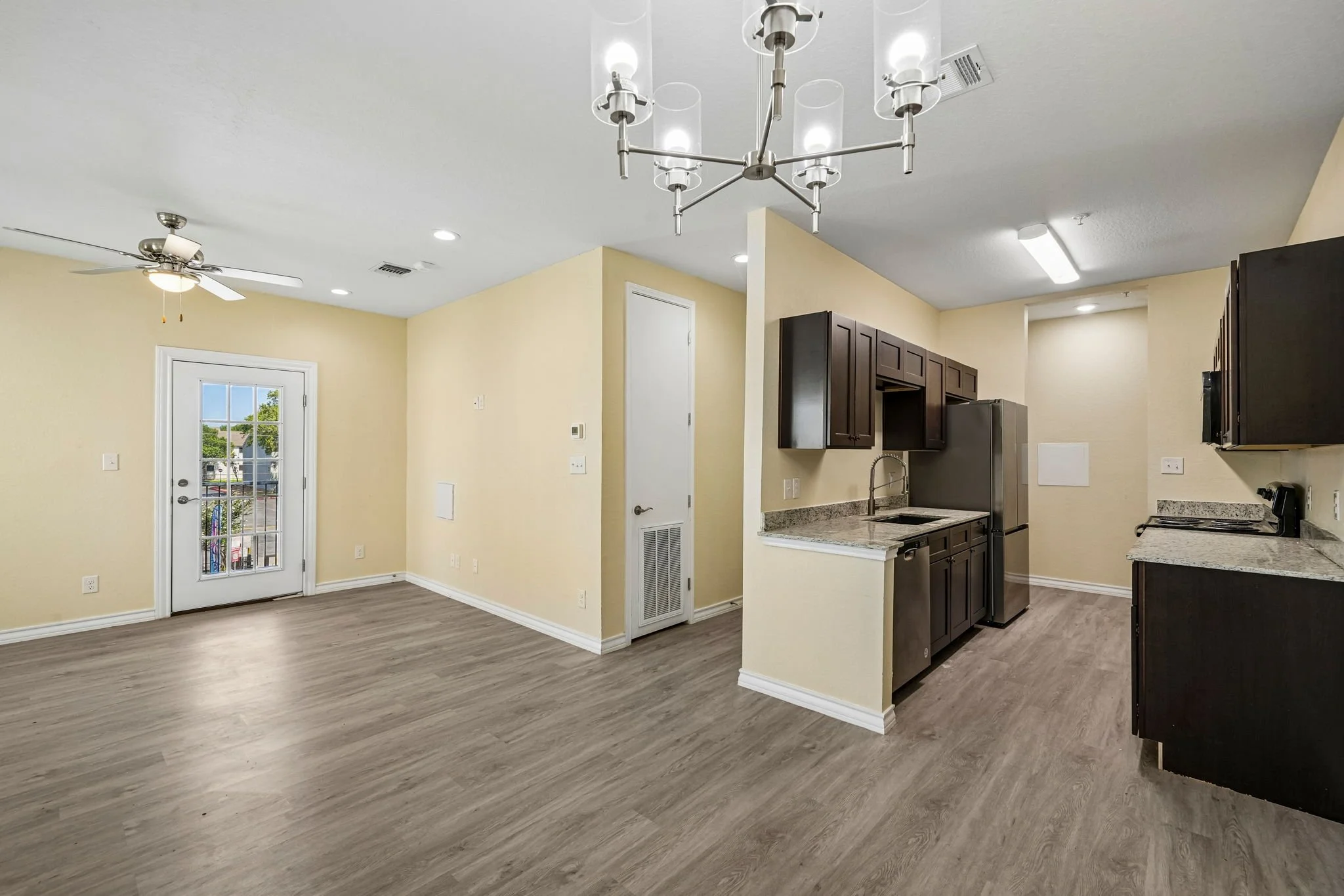 Open-concept living space with a kitchen, ceiling fan, and a door leading outside, featuring wood-look flooring and beige walls.
