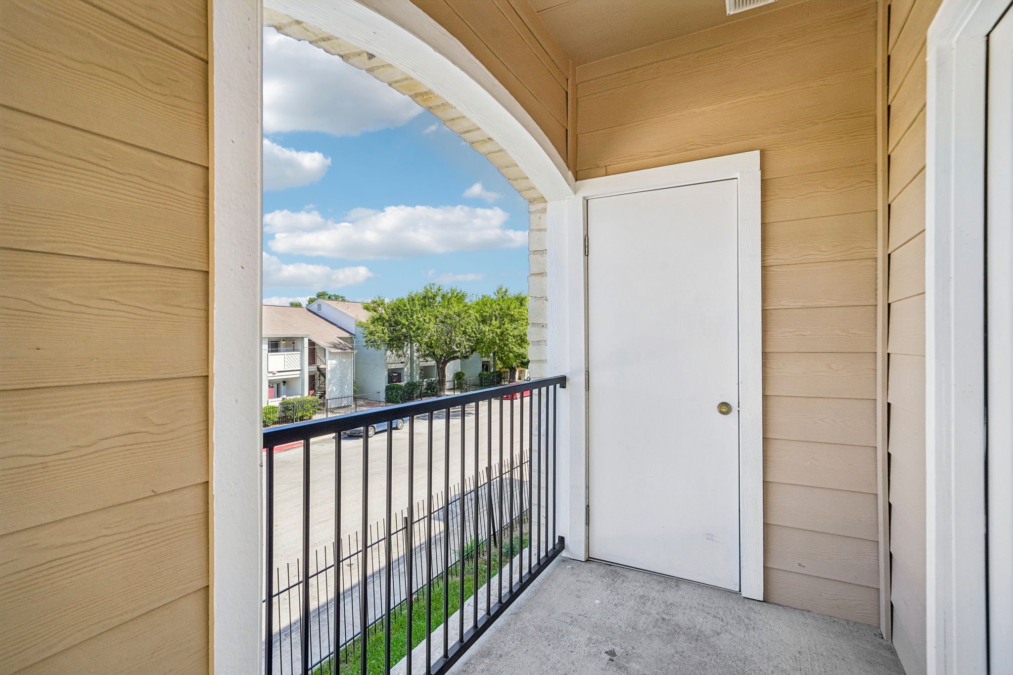 A balcony with beige horizontal siding, a white door, black metal railing, and a view of neighboring apartment buildings, trees, and a partly cloudy sky.