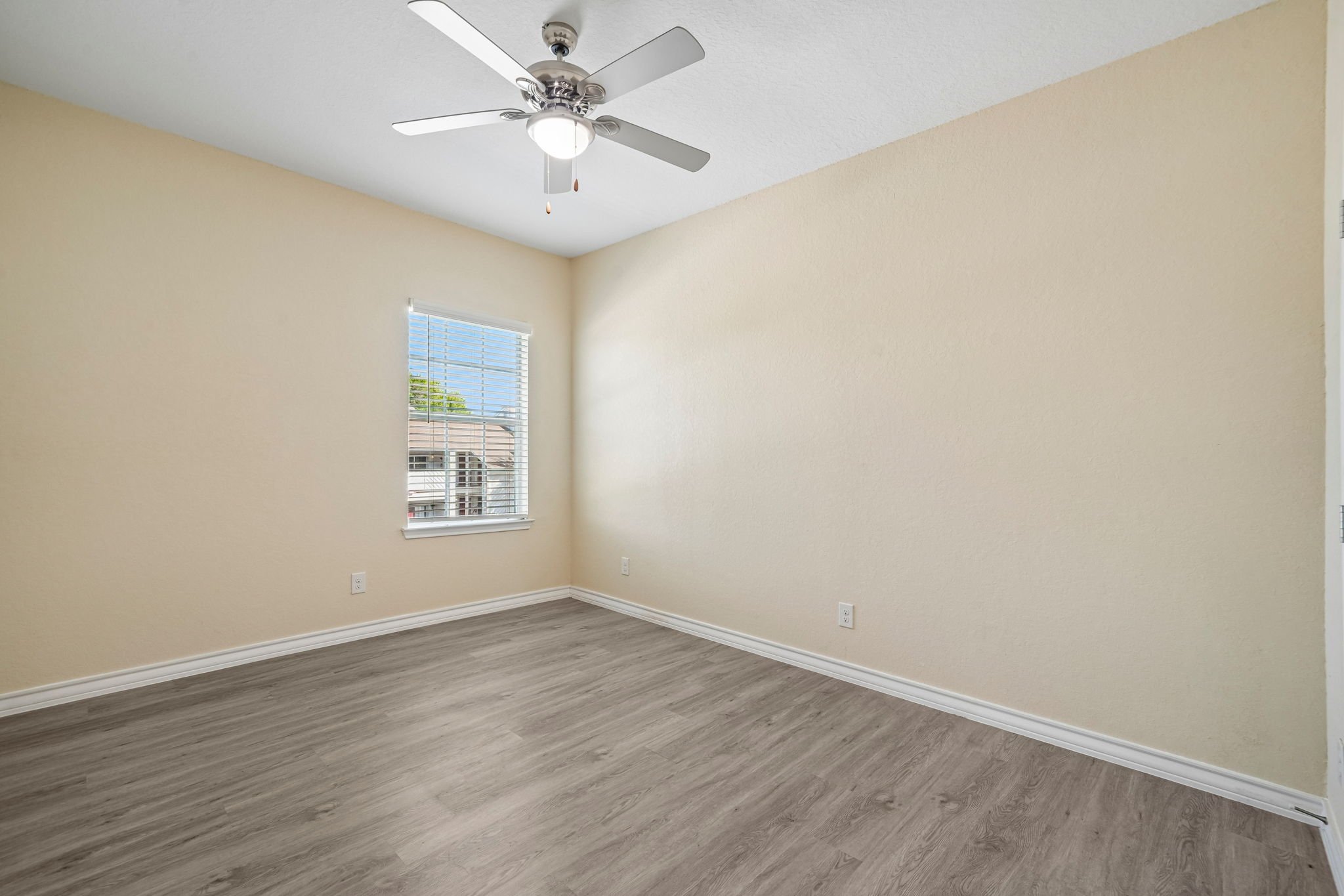 Empty bedroom with beige walls, gray wood flooring, a window with blinds, and a ceiling fan with a light.