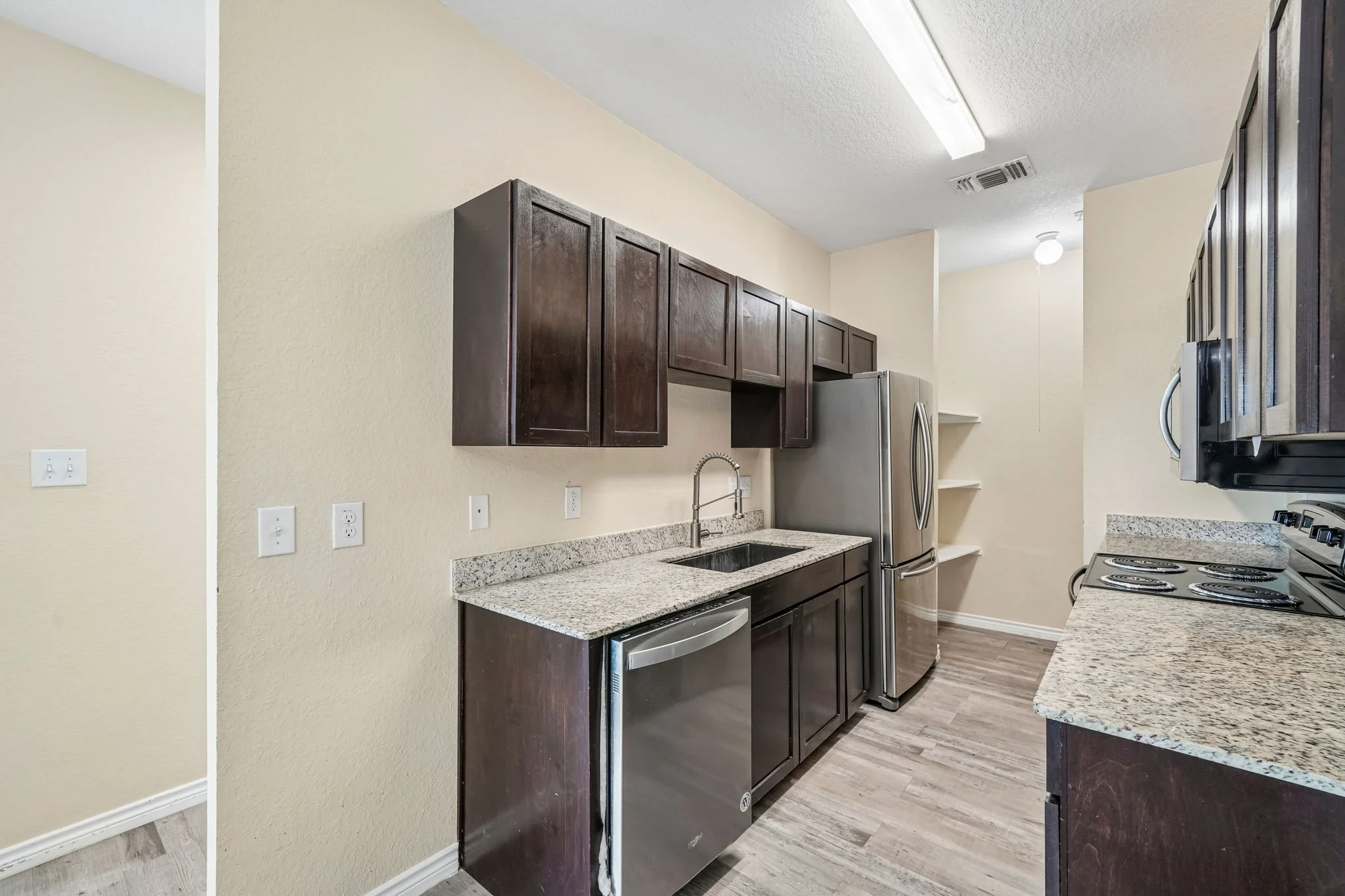 Kitchen with beige walls, dark wood cabinets, granite countertops, stainless steel refrigerator, oven, and small sections of open shelves, with electrical outlets on the wall and wood flooring.