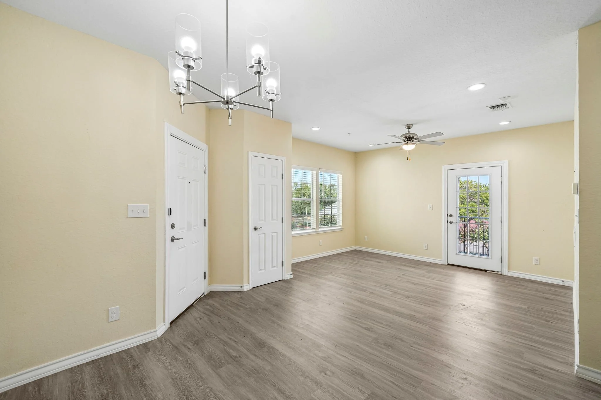 Empty living area with beige walls, wood flooring, large windows, a ceiling fan, and a modern chandelier.