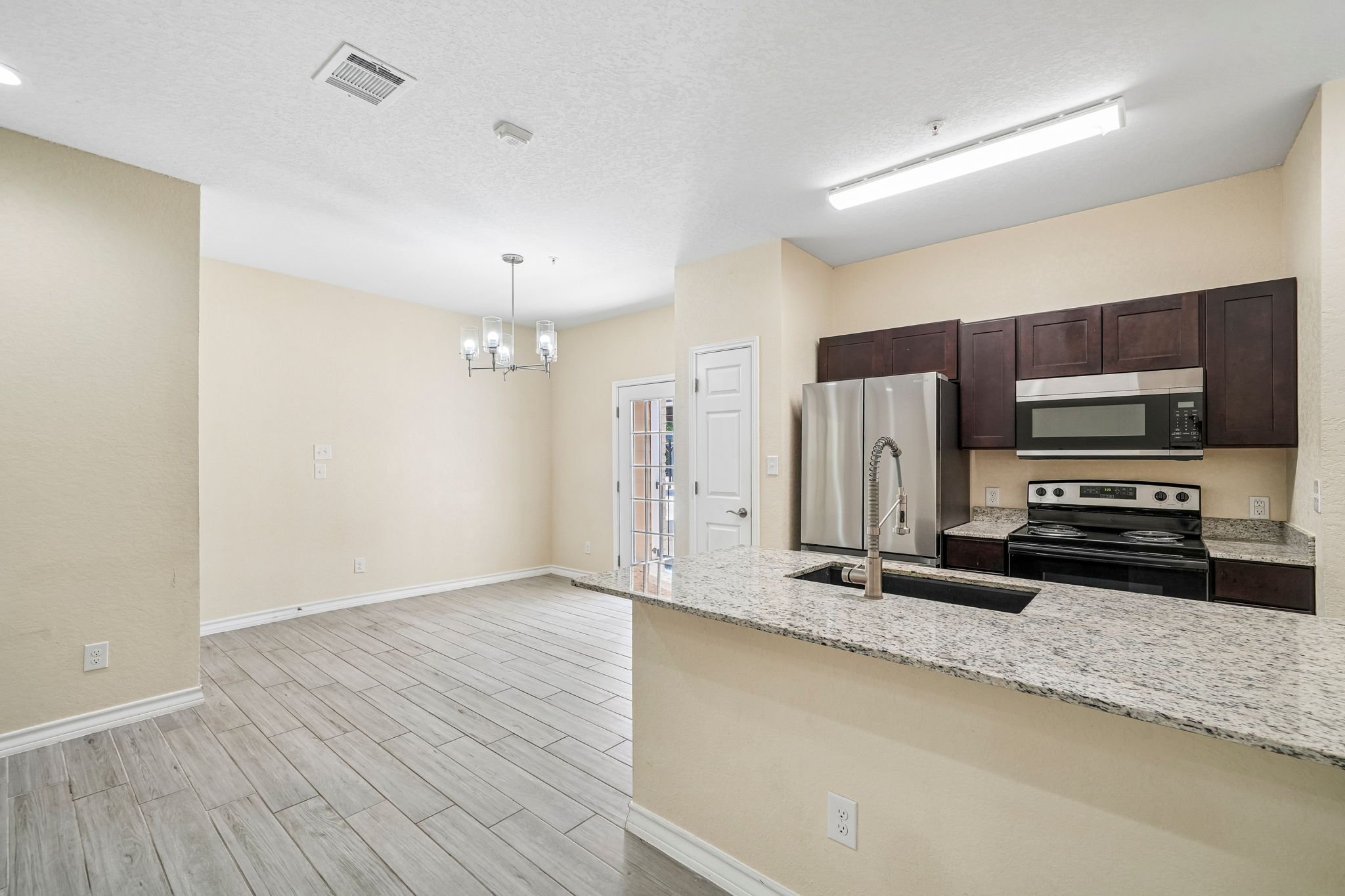 Interior view of a modern kitchen and dining area with light-colored walls, wood-like flooring, a granite countertop, stainless steel appliances including a refrigerator and microwave, dark wood cabinets, and a ceiling light fixture.