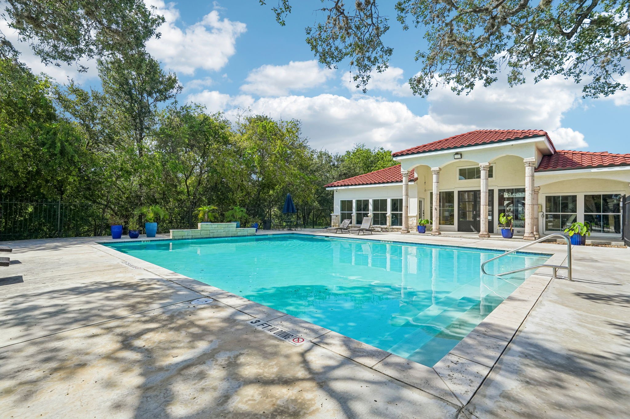 A swimming pool with a clubhouse at La Marquesa Apartments featuring white walls, a red tile roof, and large glass windows. The area around the pool has lounge chairs, potted plants, and a shaded umbrella. San Antonio ,Texas.