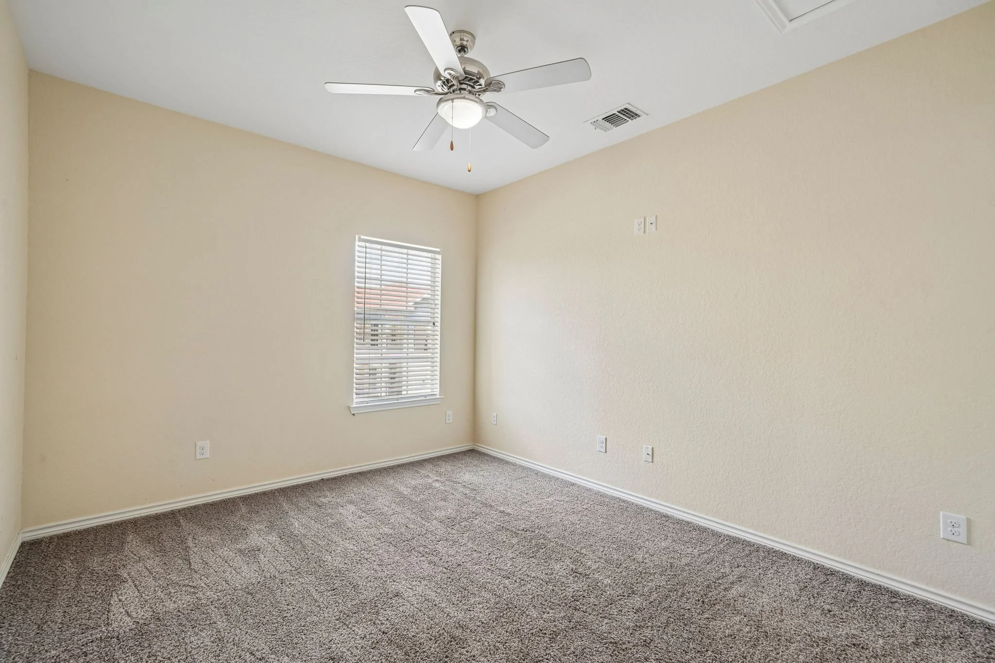 Empty bedroom with beige walls, brown carpeted floor, a window with blinds, and a ceiling fan with lights.