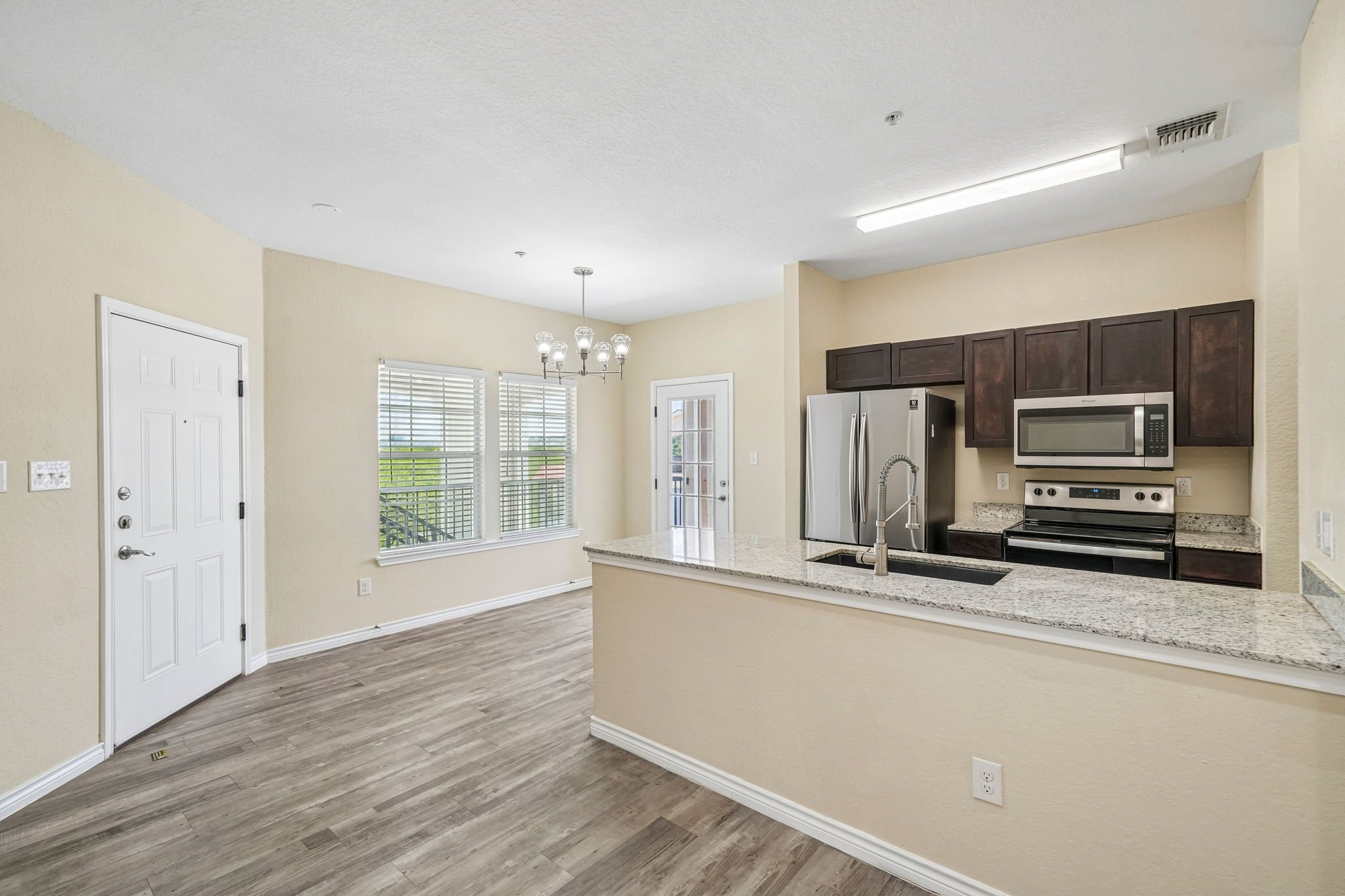 Interior view of a modern kitchen with granite countertops, dark cabinets, stainless steel refrigerator and microwave, sunlight through large windows, and a chandelier over the dining area.