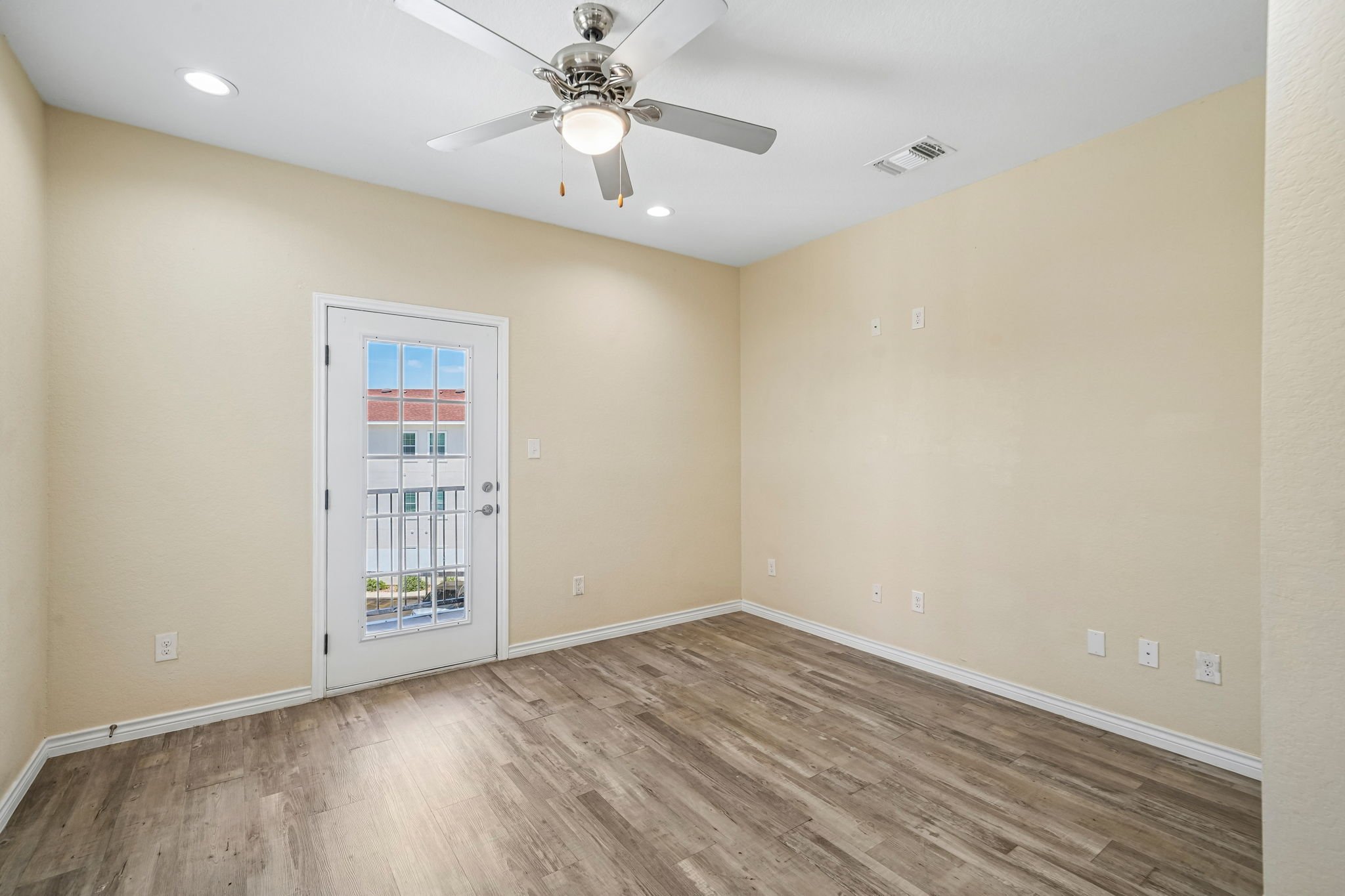 Empty living room with beige walls, a white ceiling fan, hardwood flooring, a door with a window, and multiple electrical outlets and switches.