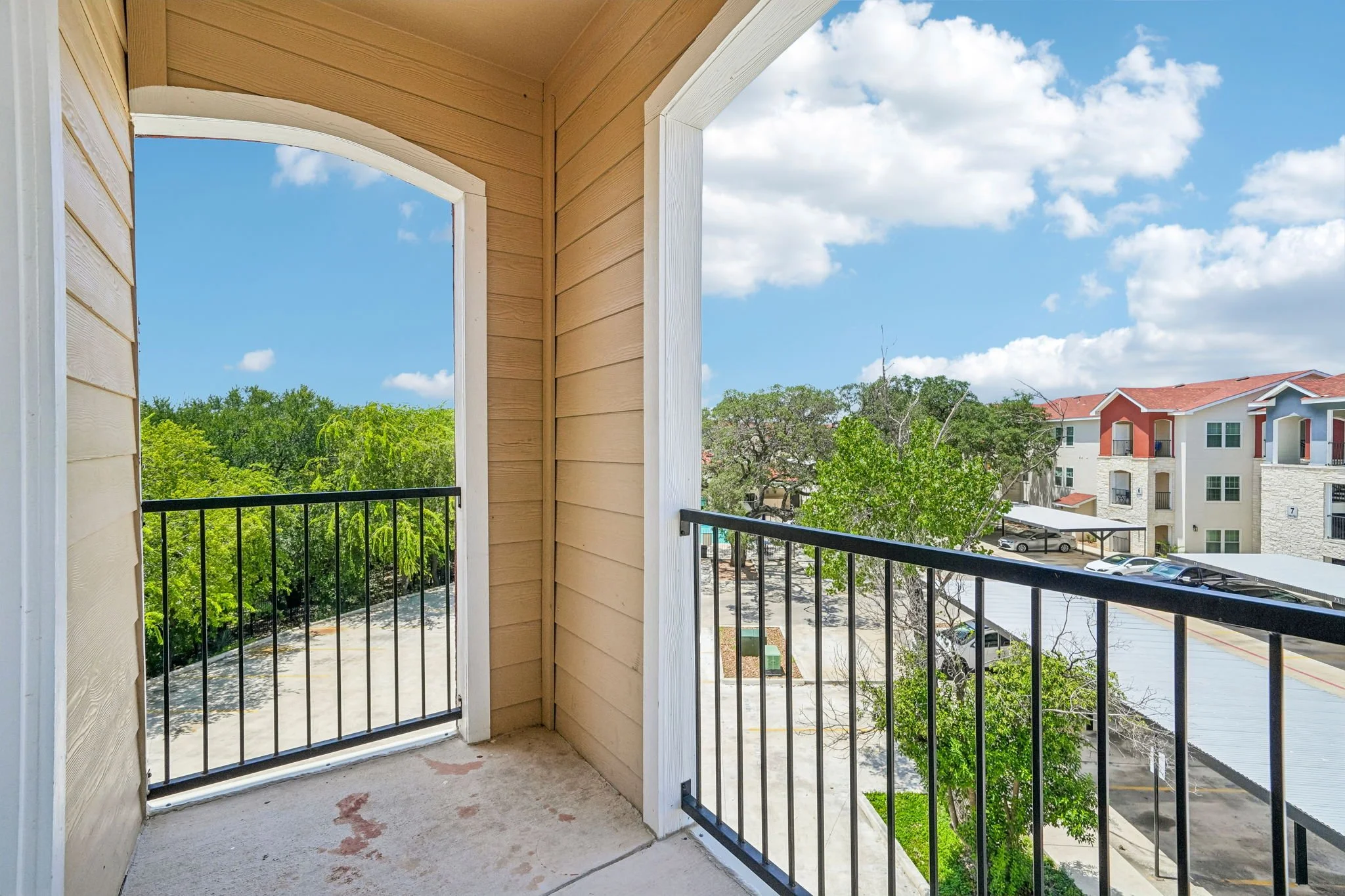 View from a balcony at La Marquesa Apartments showing a tree-lined area, parking lot, and neighboring buildings with red and white exteriors under a partly cloudy sky.