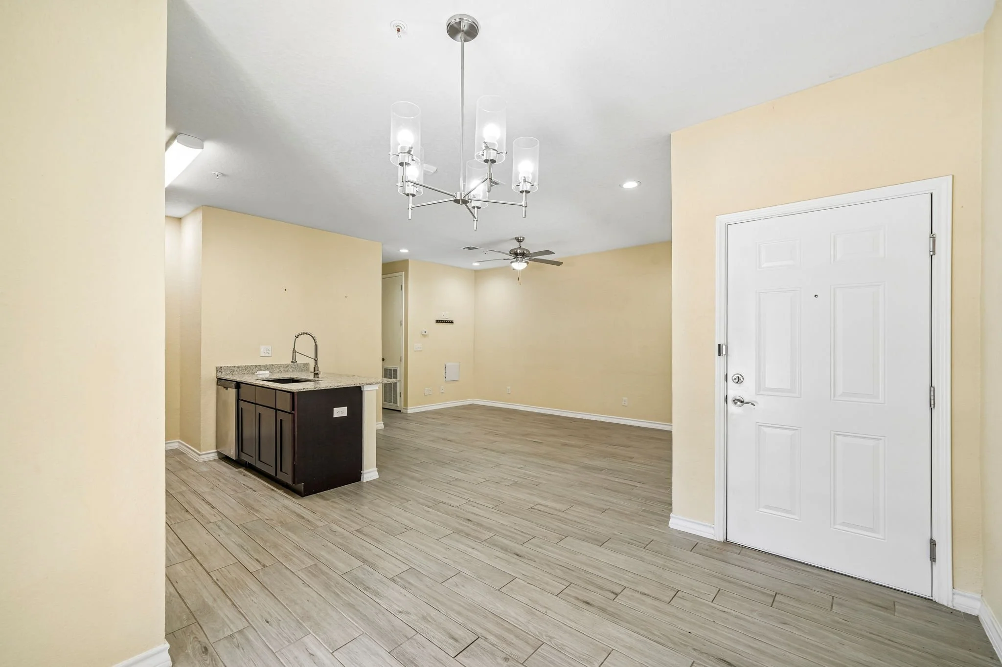 Empty living area with light-colored walls, wood-look tile flooring, a ceiling fan, and a modern chandelier, near an entry door with a peephole.