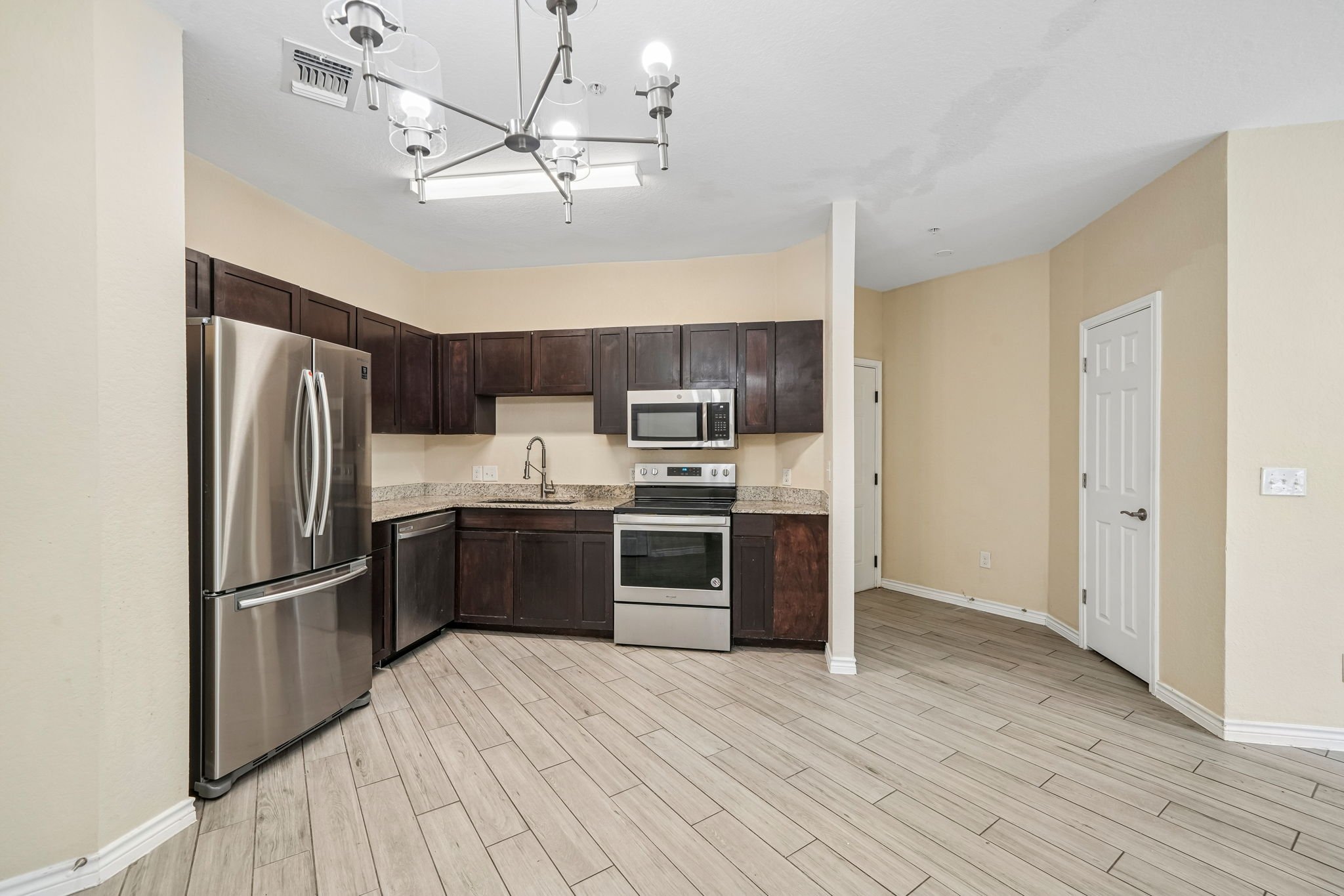 Kitchen with dark wood cabinets, stainless steel refrigerator, oven, microwave, beige walls, light wood flooring, and a ceiling light fixture.