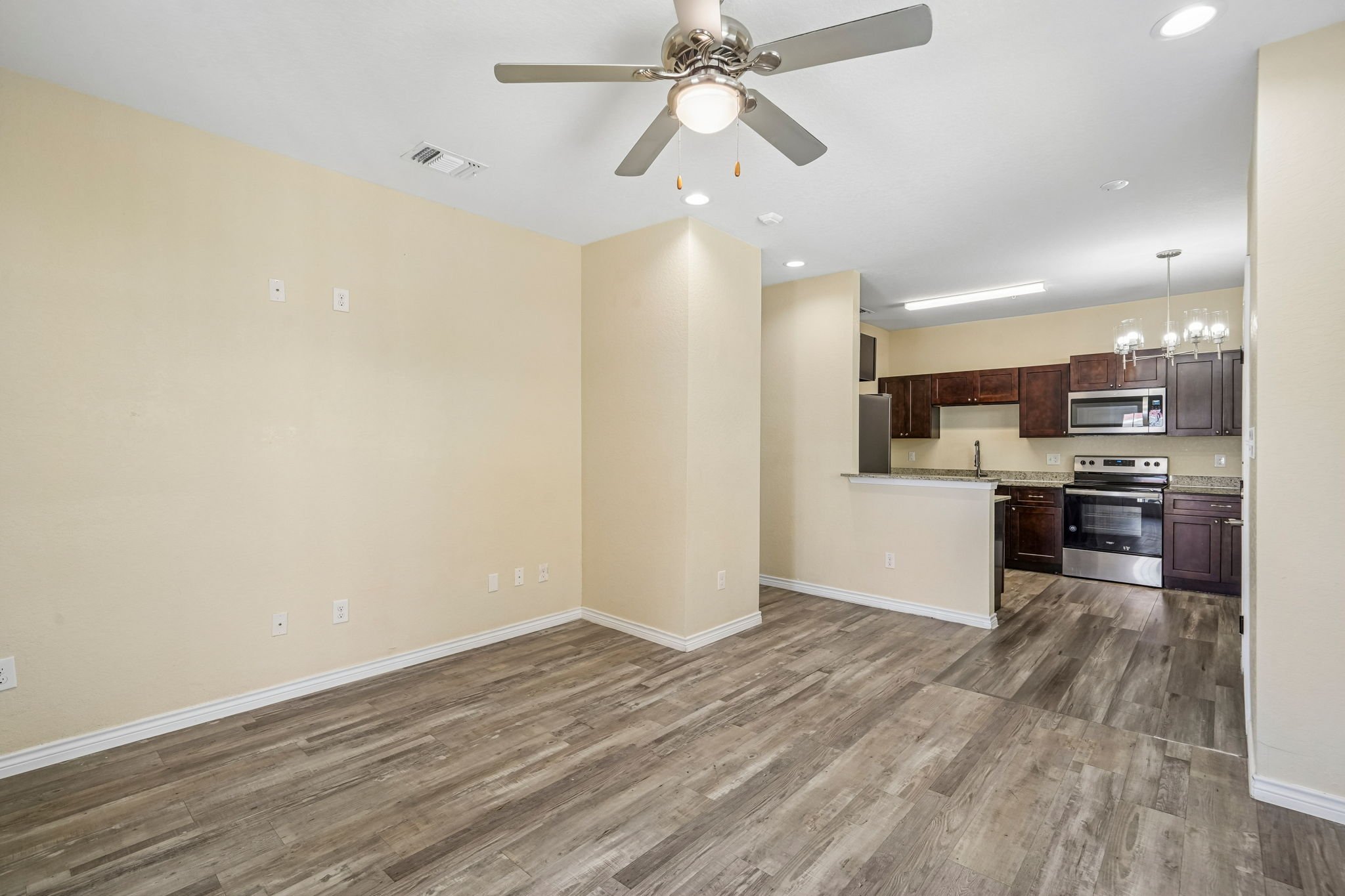 Empty living room facing open kitchen with dark wood cabinets, stainless steel appliances, and light-colored walls and flooring, with ceiling fan and lighting fixtures.