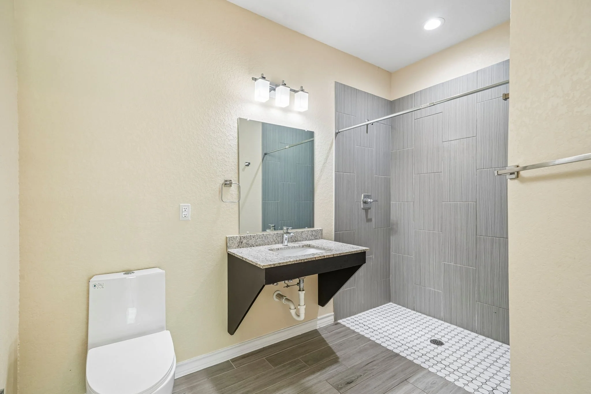 A modern bathroom with a light-colored wall, gray tile shower, small white hexagonal floor tiles in shower, wall mounted mirror, granite sink countertop, and white toilet.