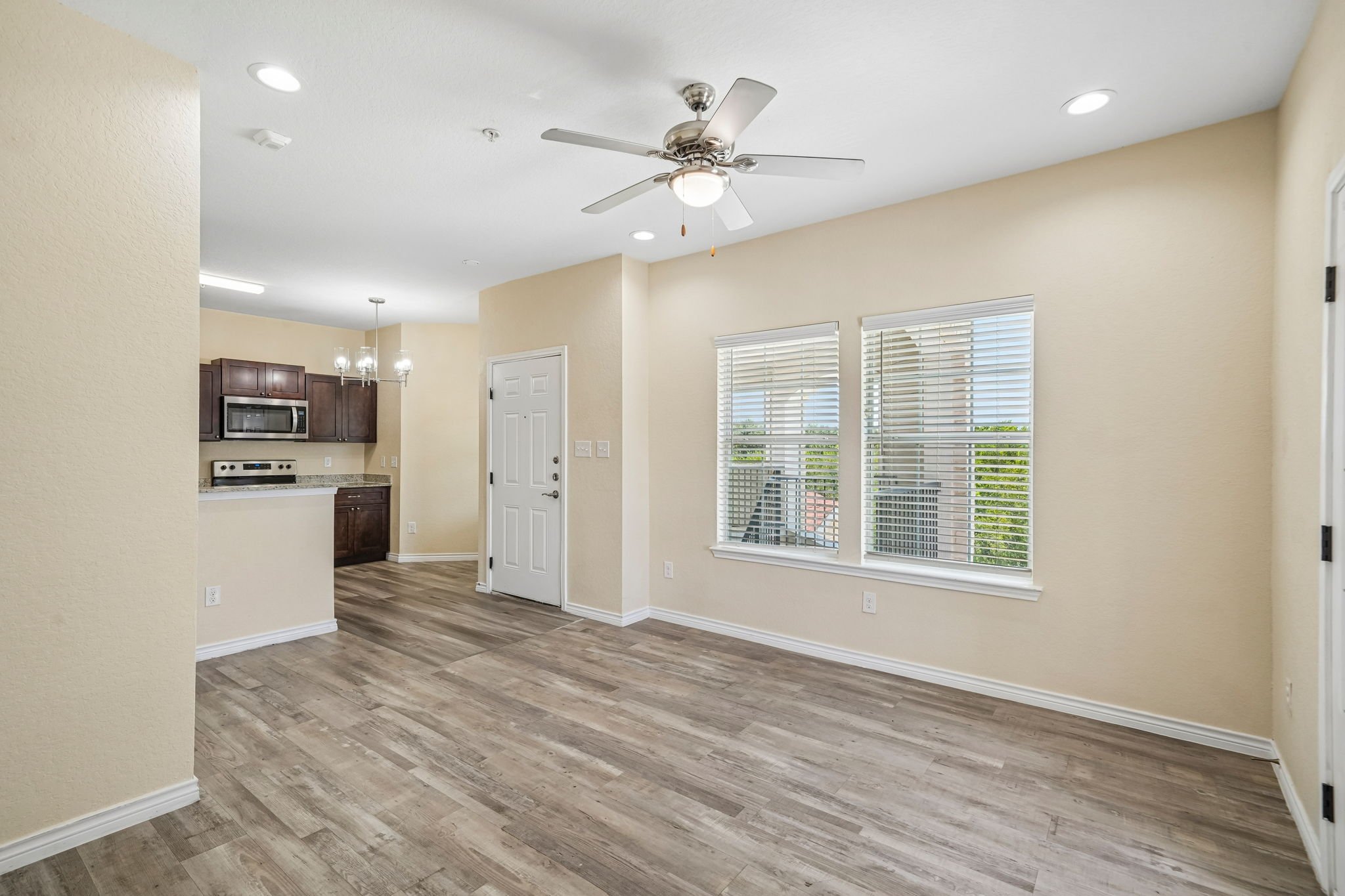 Empty living room area with beige walls, large windows with white blinds, and wood-look flooring, overlooking the kitchen with dark cabinetry and stainless steel appliances.