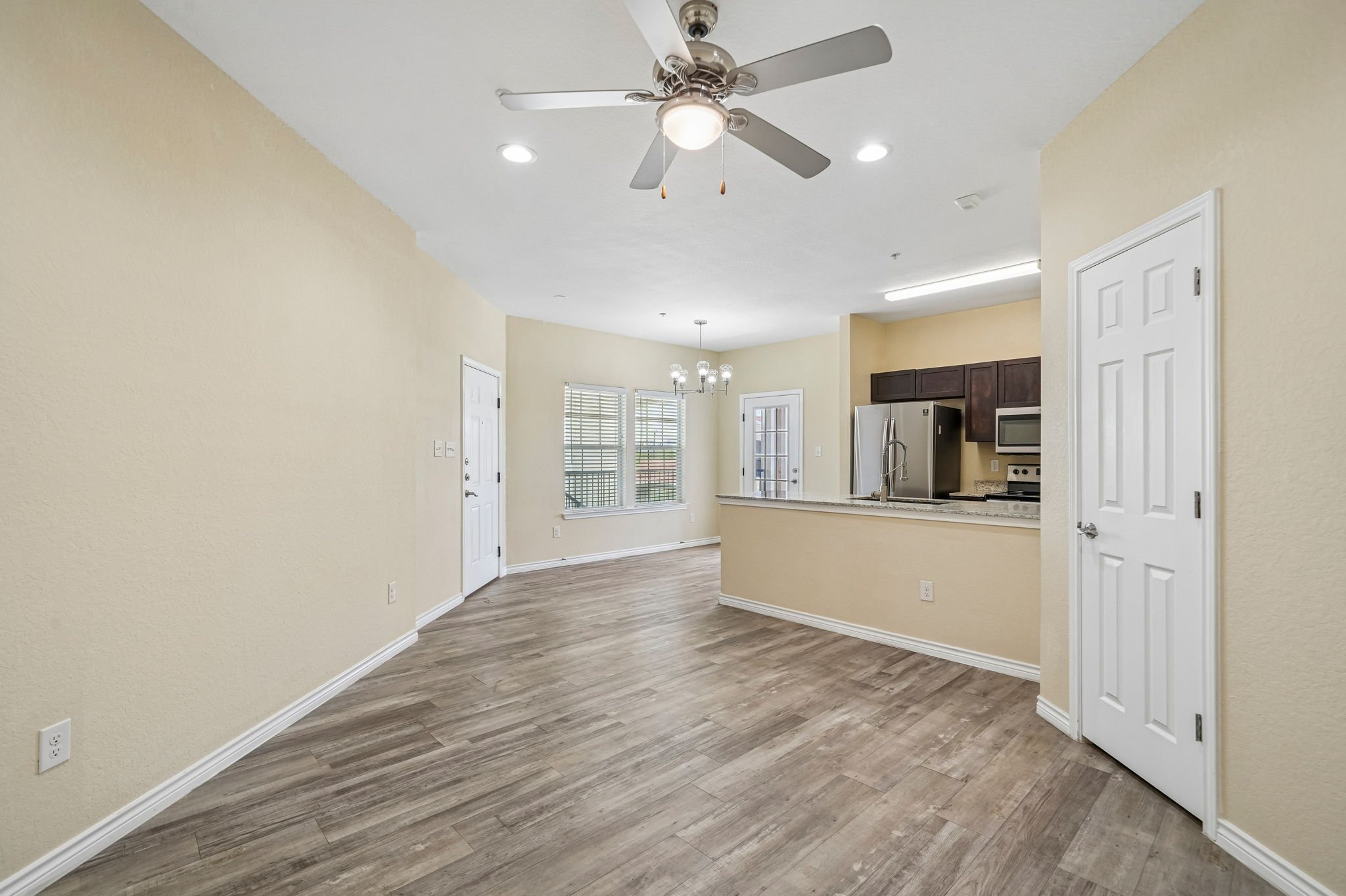 Empty living room with beige walls, wood flooring, kitchen area with stainless steel refrigerator, microwave, and dark wood cabinets, white doors, large windows with blinds, ceiling fan, and chandelier lighting.