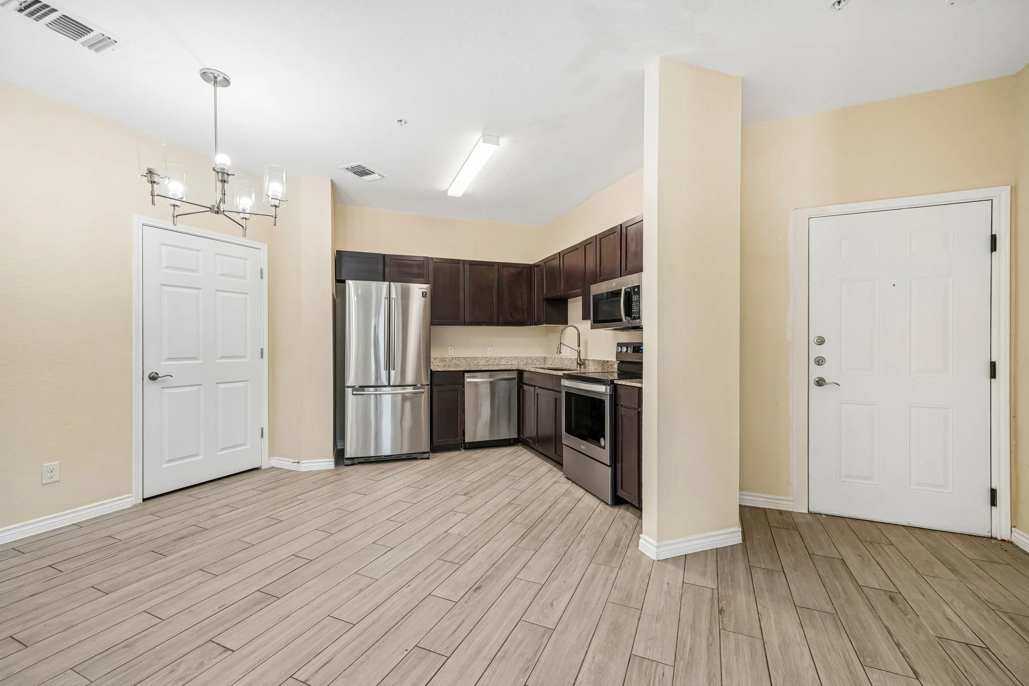 Empty kitchen with beige walls, light wood flooring, dark wood cabinets, stainless steel refrigerator, microwave, oven, and a white door, with a chandelier and a fluorescent ceiling light.