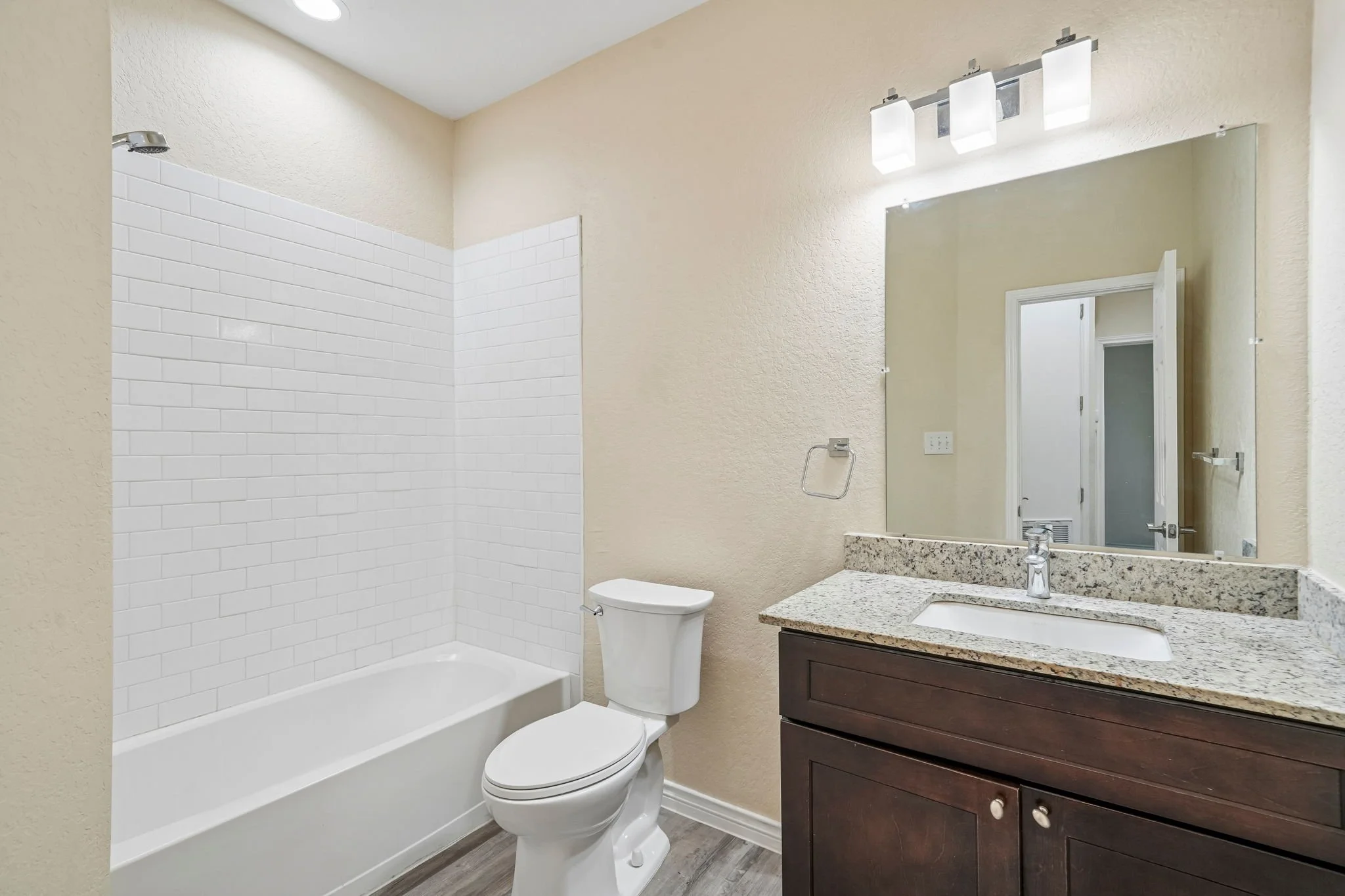 Bathroom with bathtub, white subway tiles, toilet, granite countertop vanity, mirror, light fixture, and door.