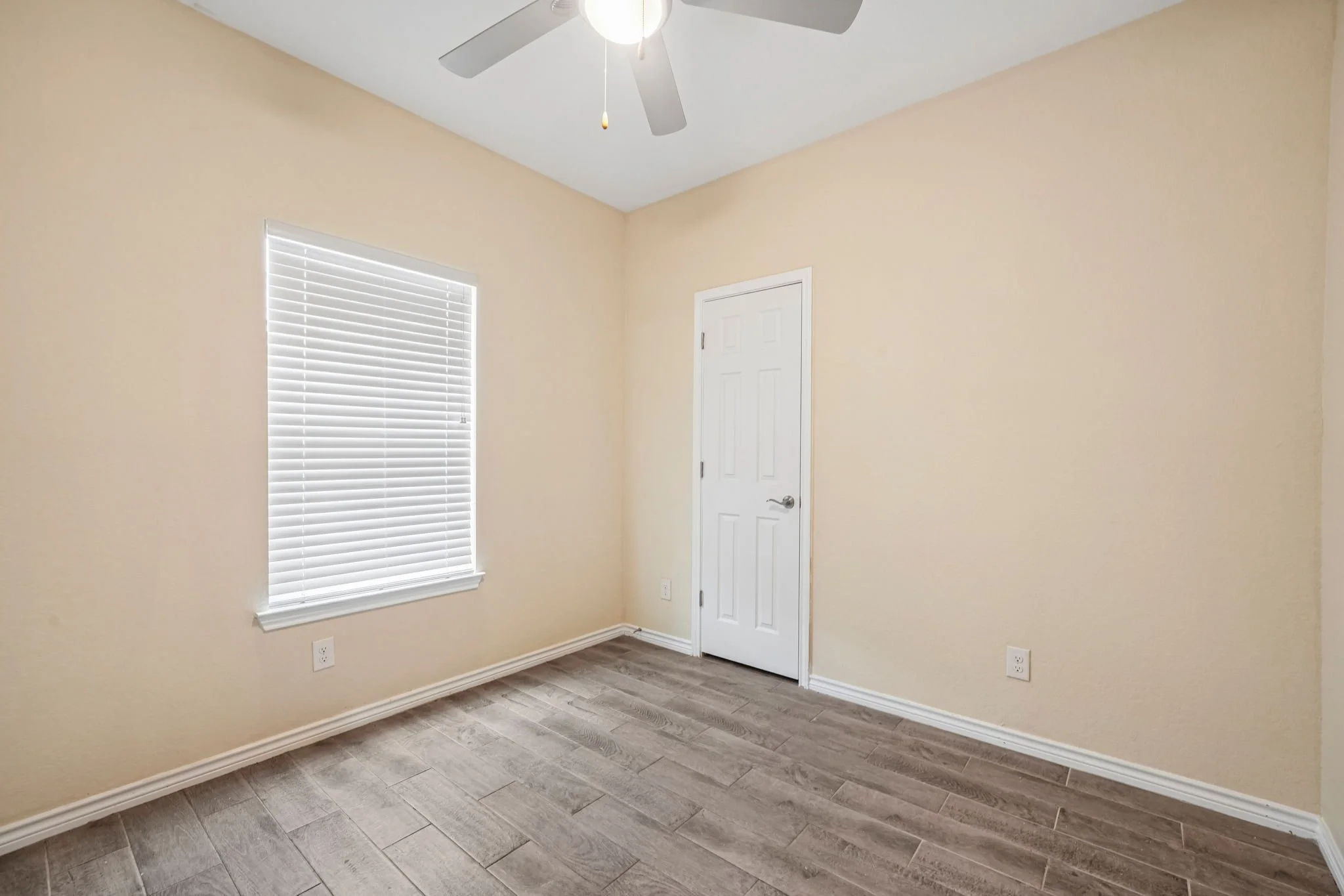 Empty bedroom with beige walls, a white door, a window with white blinds, gray wood-like floor, and a ceiling fan.