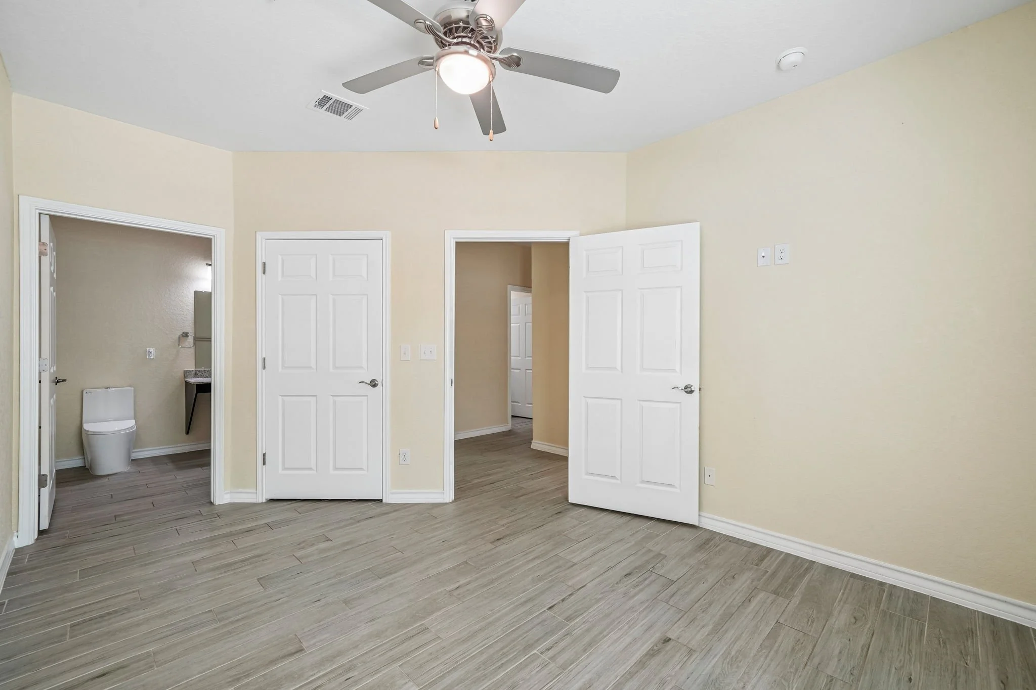 Empty primary bedroom with light-colored walls, multiple doors, a ceiling fan with a light, and wood-patterned flooring.