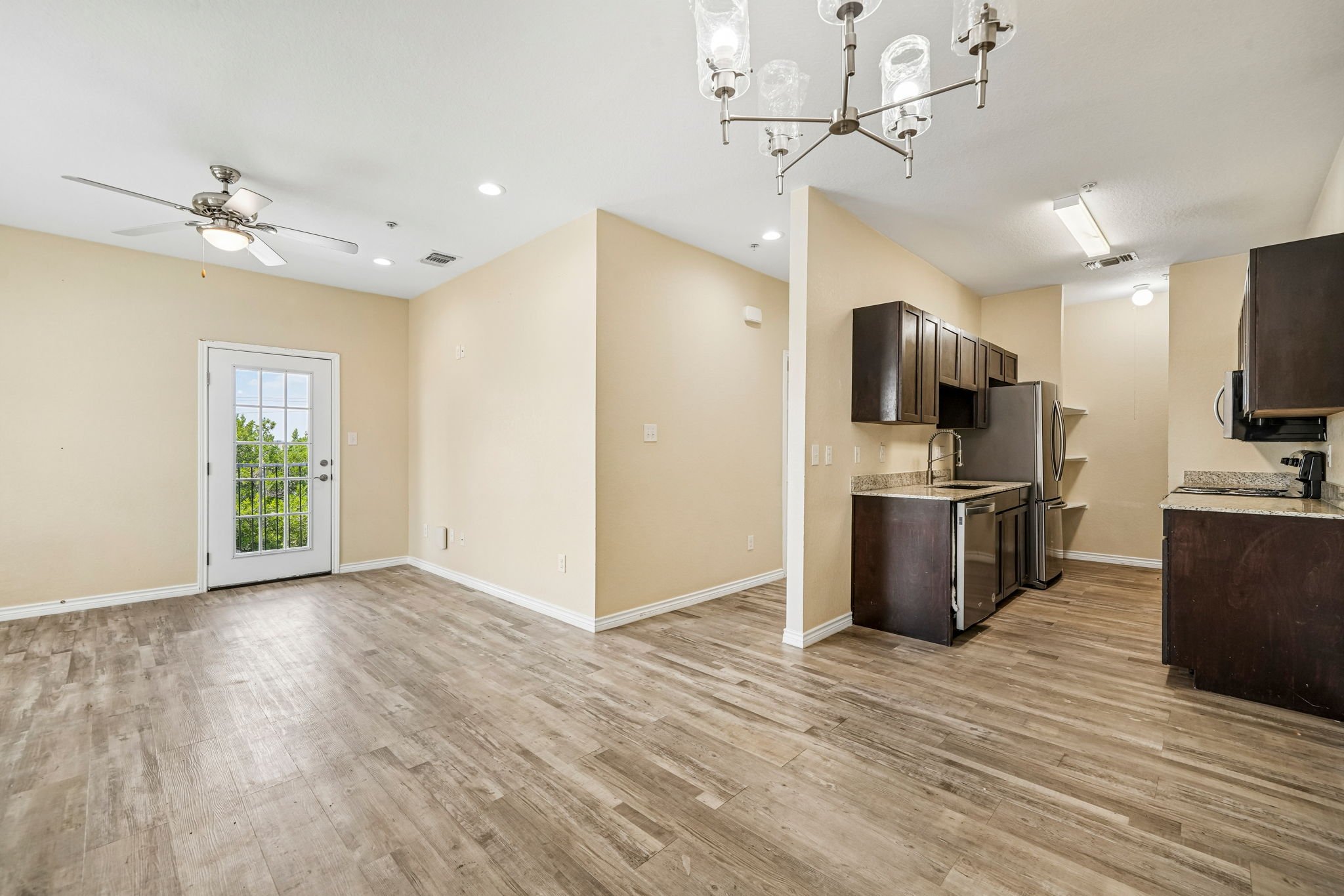 Empty living room and kitchen area with beige walls, wood flooring, ceiling fans, and a door with a window leading outside. Kitchen features dark wood cabinets, stainless steel refrigerator, microwave, and granite countertops.
