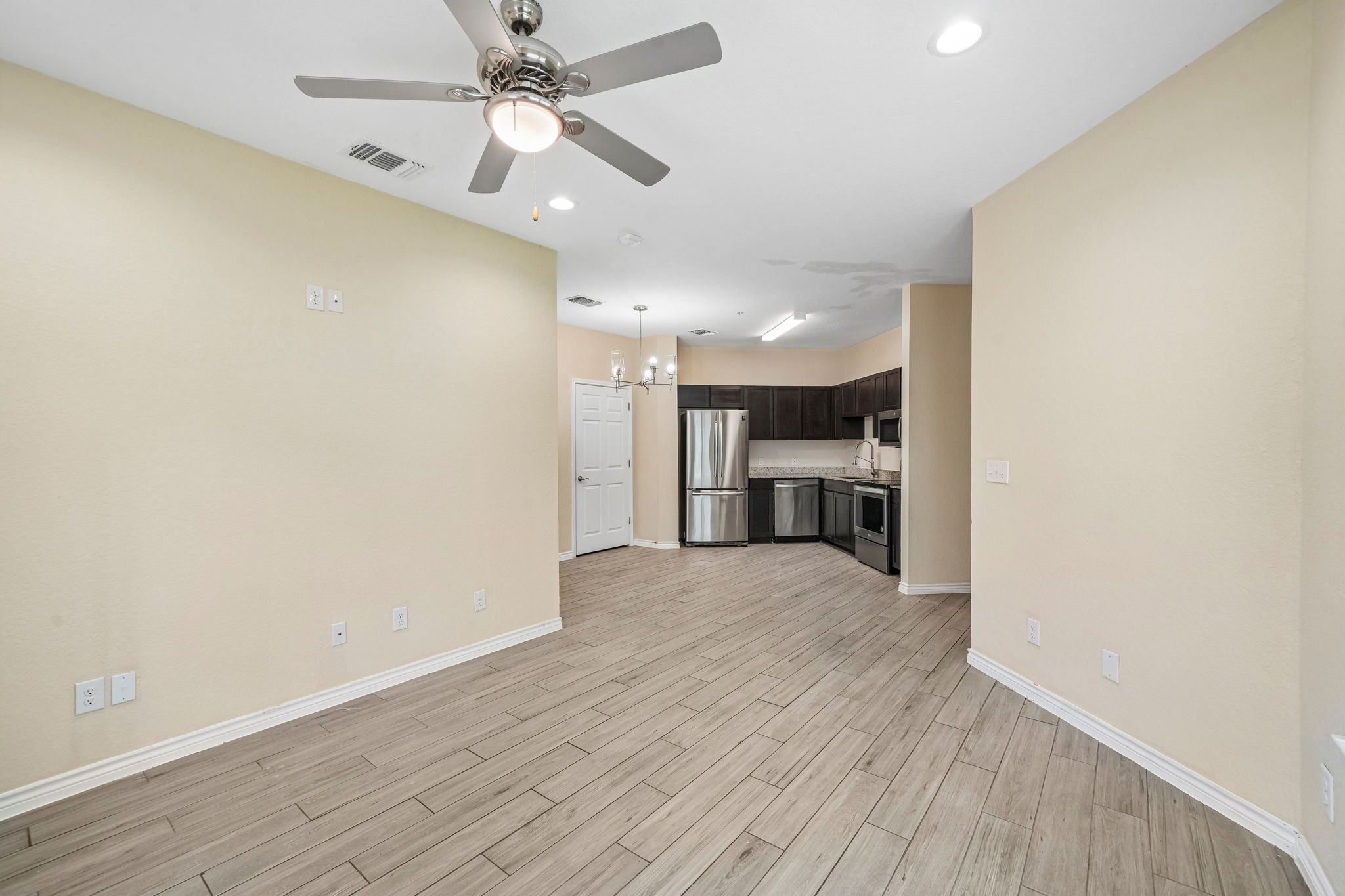 Empty living space with beige walls, wood-look flooring, kitchen in background, and ceiling fan.