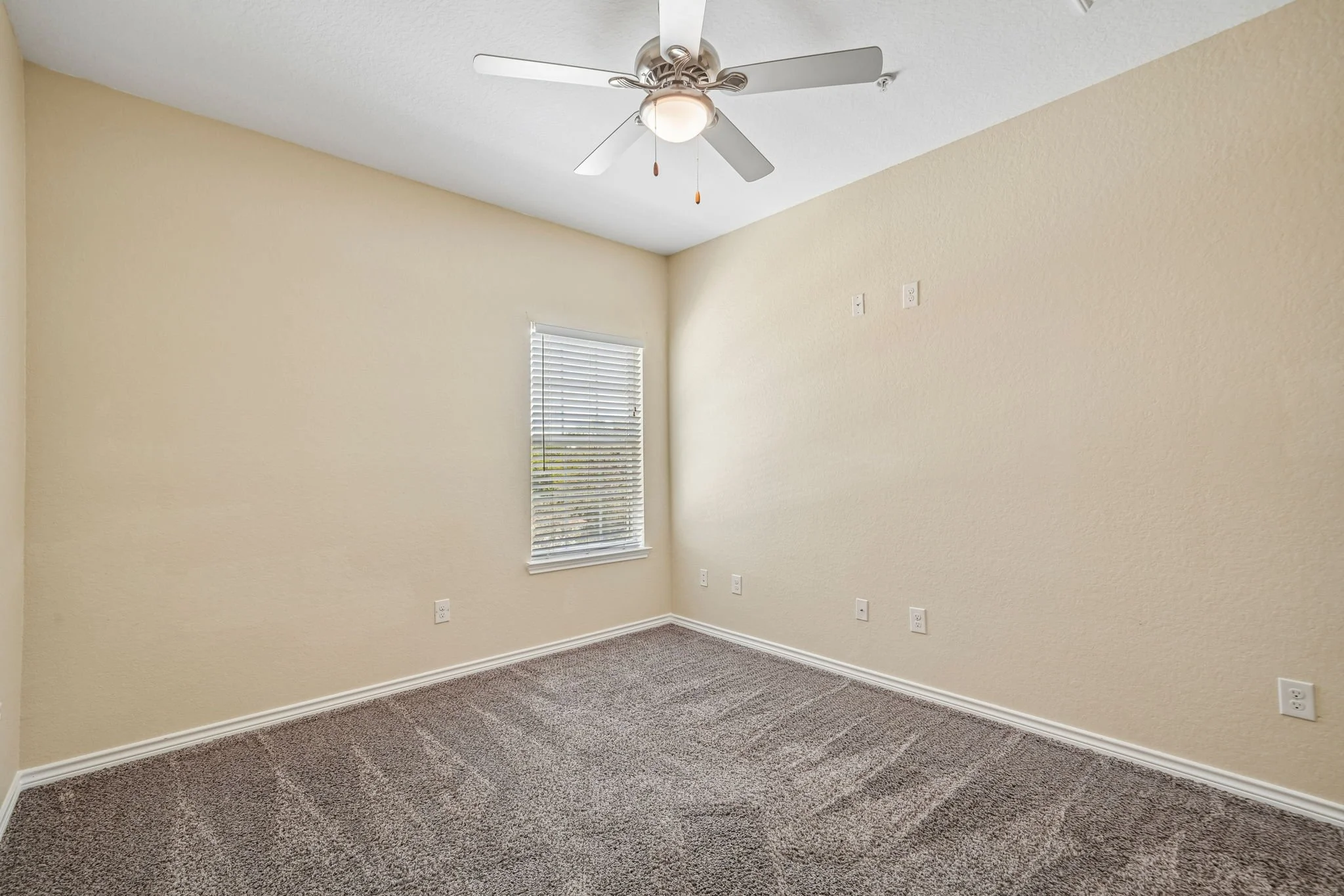 Empty bedroom with beige walls, a white ceiling fan with light, a window with blinds, and multiple electrical outlets.