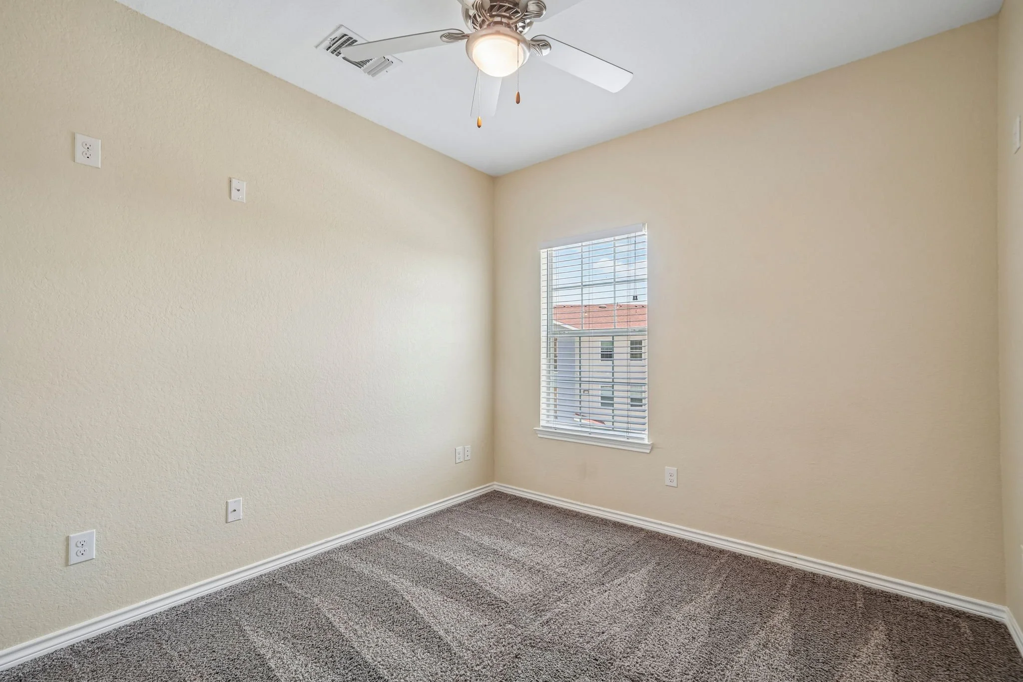 Empty bedroom with beige walls, a large window with blinds, ceiling fan with a light, and electrical outlets along the walls, carpeted floor.