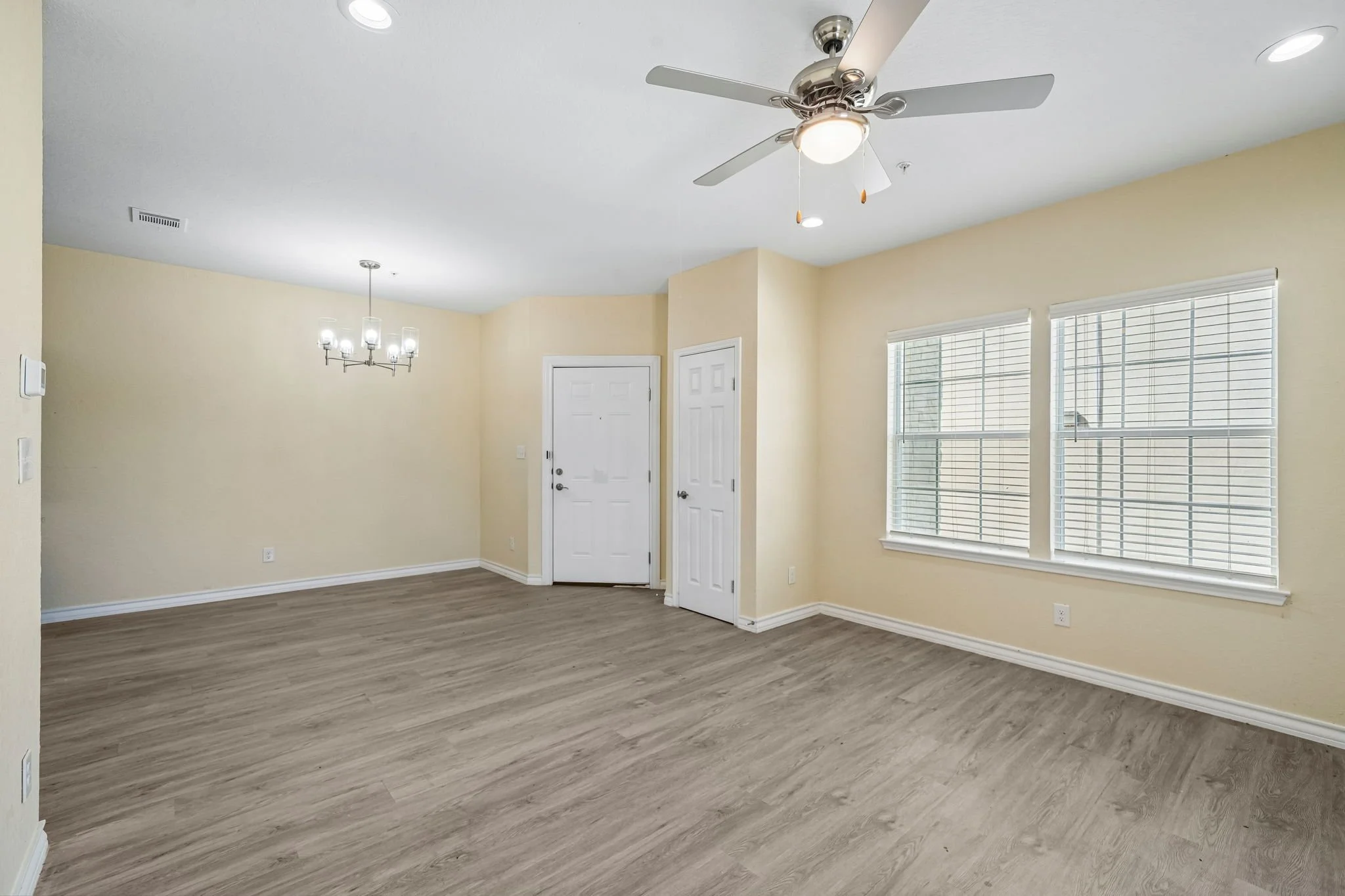Empty living room with beige walls, wooden flooring, large windows with blinds, a ceiling fan, and a chandelier.