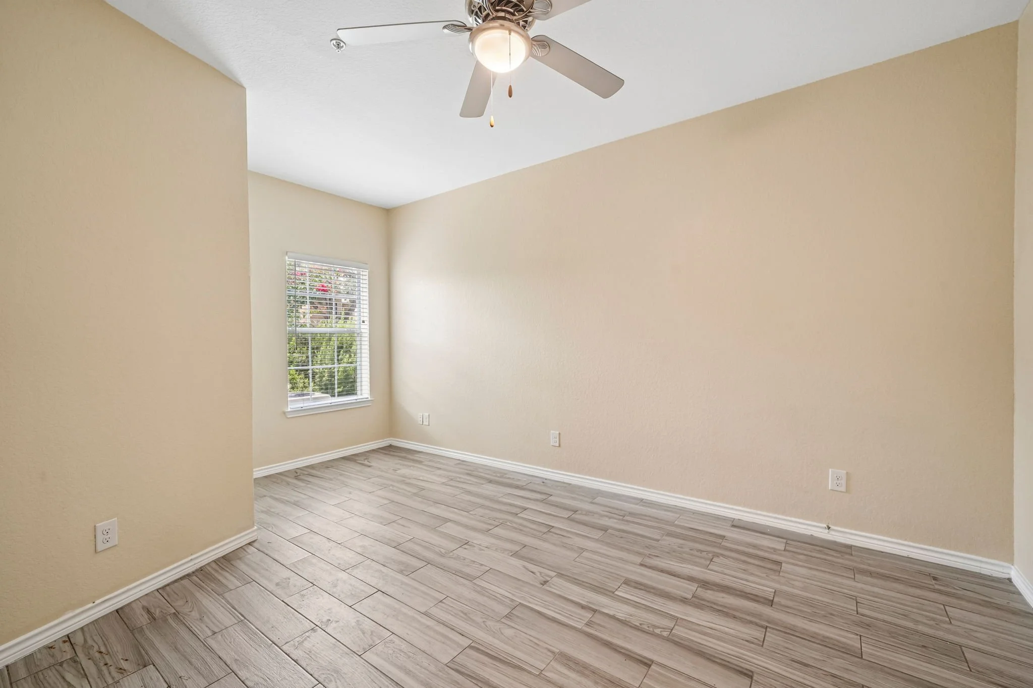 Empty primary bedroom with beige walls, a window with blinds, a ceiling fan with a light, and wood-look flooring.
