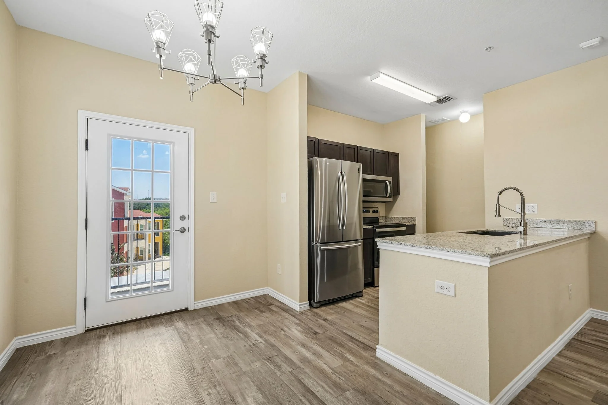 Interior view of a modern kitchen with beige walls, a granite countertop, stainless steel refrigerator, microwave, dark wood cabinets, wood flooring, a white door with glass panels leading outside, and a contemporary chandelier overhead.