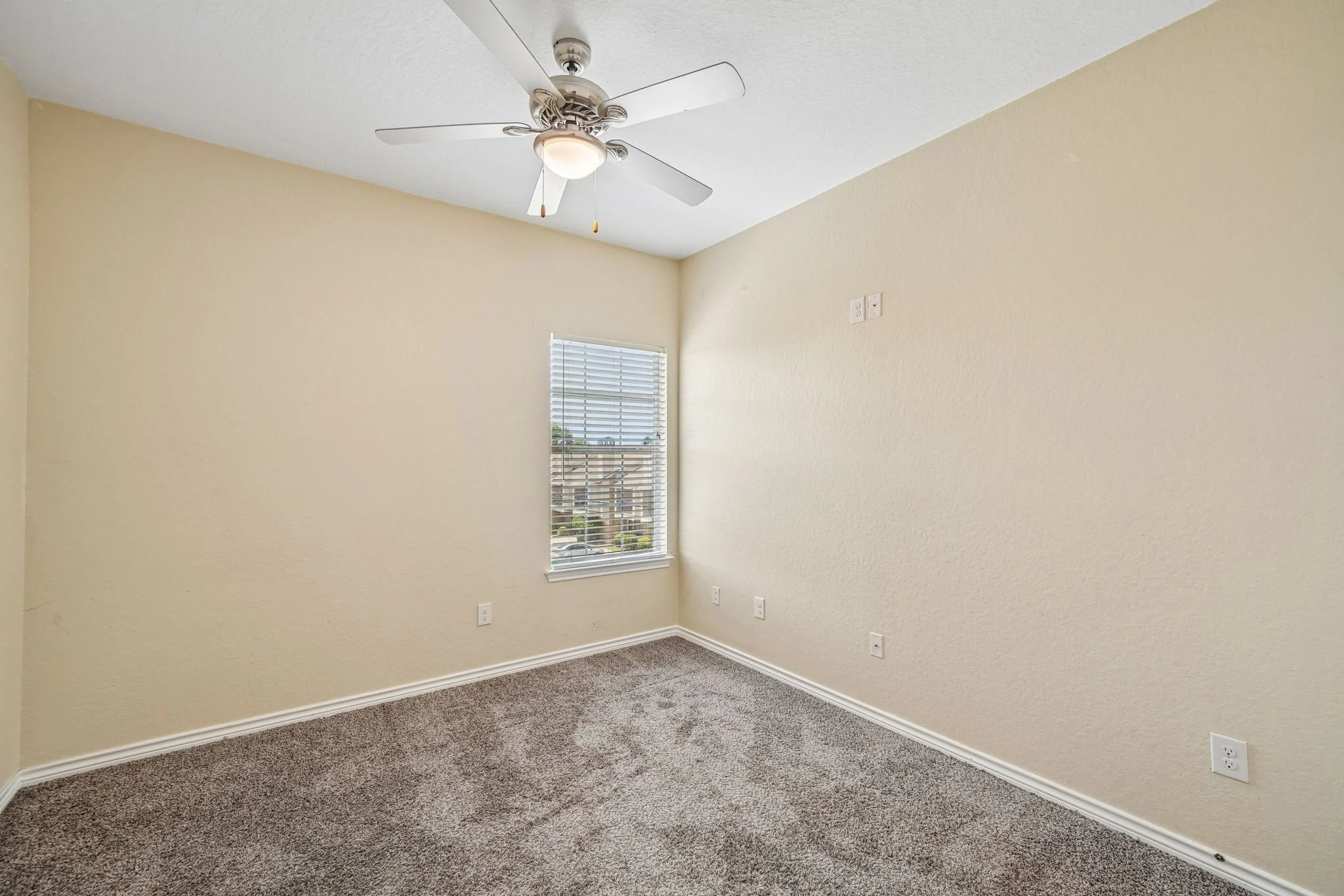 Empty bedroom with beige walls, carpeted floor, a ceiling fan with light, a window with blinds, and multiple electrical outlets.