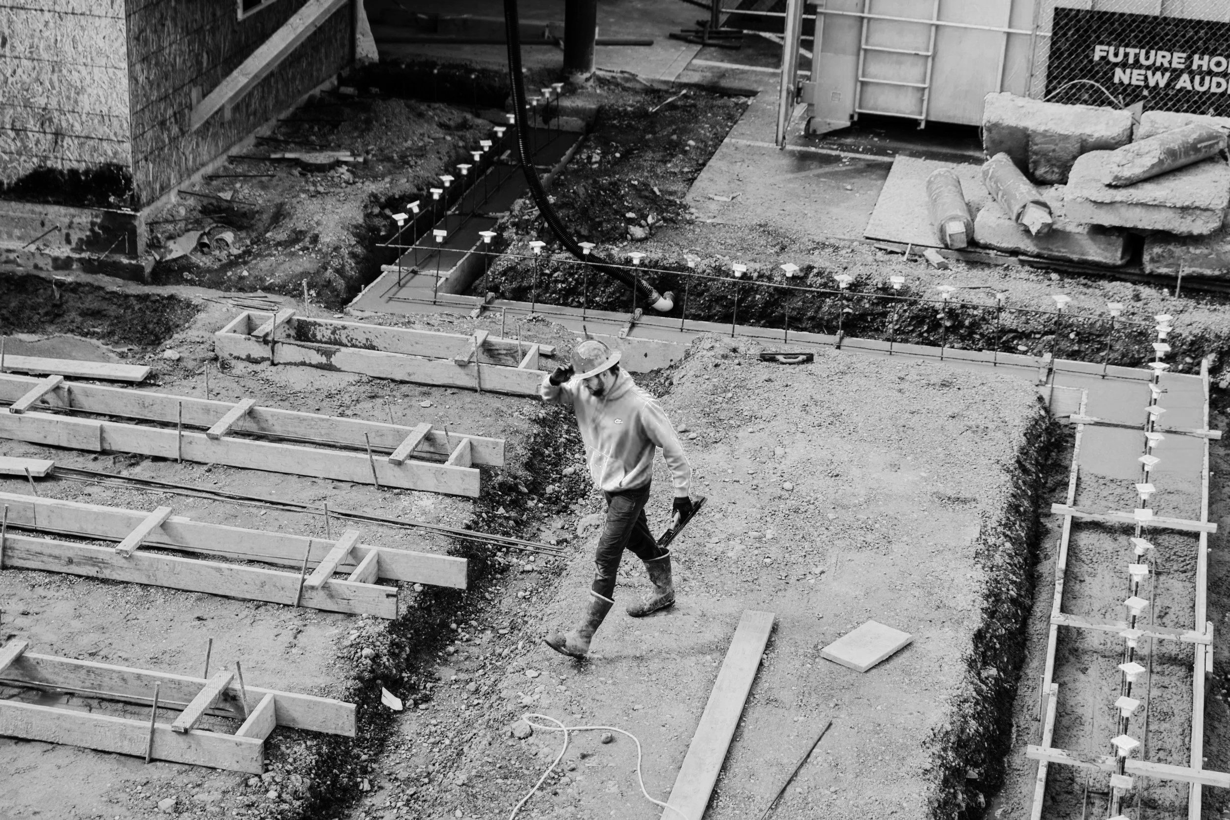 Construction site with a worker wearing a hard hat standing near a foundation outline, with wooden forms and rebar, and dirt and tools surrounding the area.