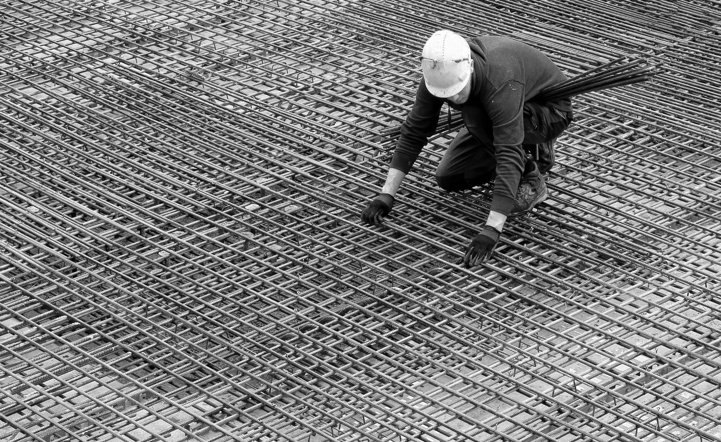 Construction worker in a hard hat crouching on a steel rebar framework at a building site.