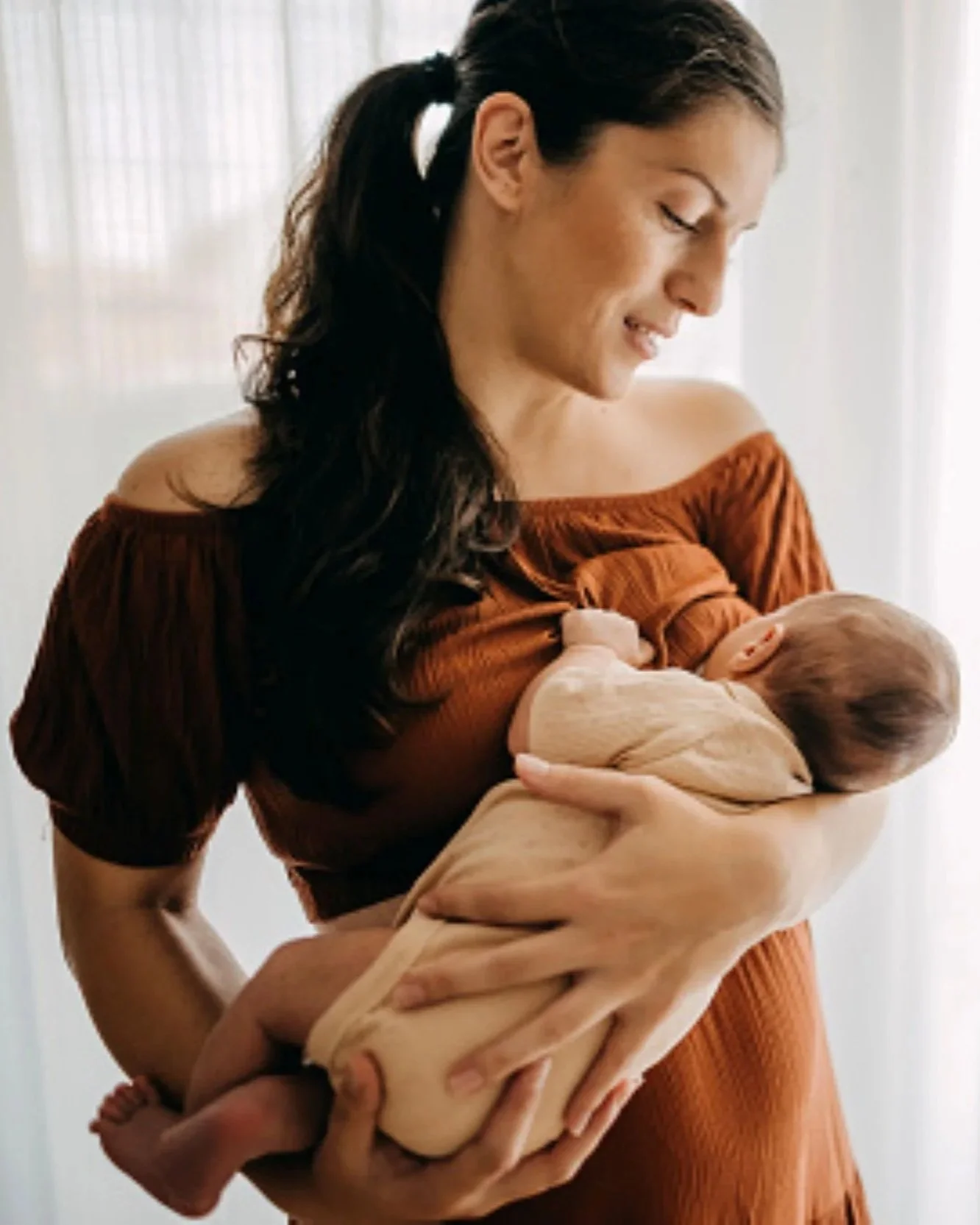 A woman with long dark hair in a ponytail holding a baby wrapped in a beige blanket near her chest, standing against a light background. She is wearing an off-your-shoulder rust-colored dress and looking down at the baby with a gentle expression.