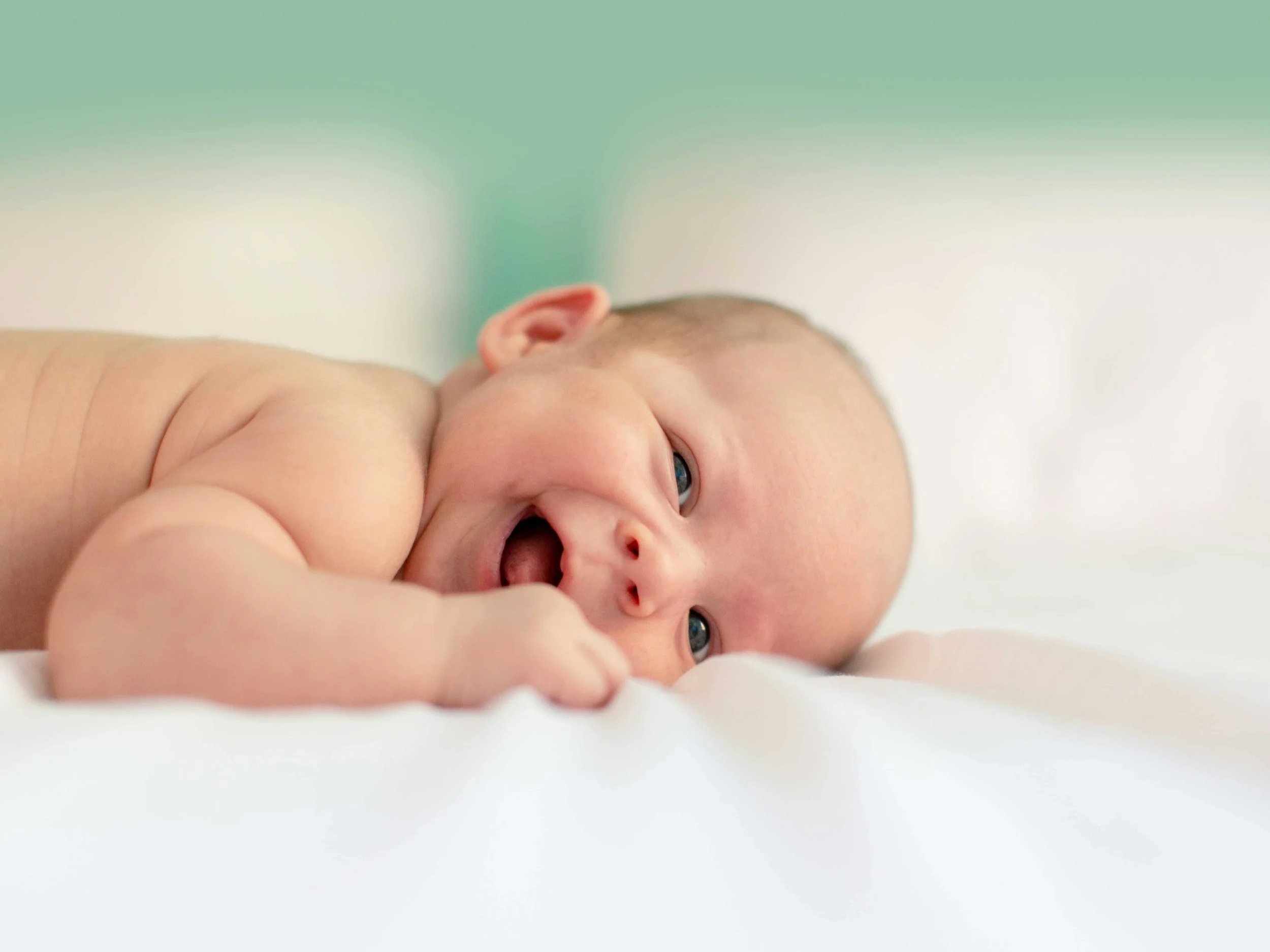 Close-up of a newborn baby lying on their stomach on a soft surface, with wide open eyes, mouth open, and making a playful face, against a blurred pastel background.