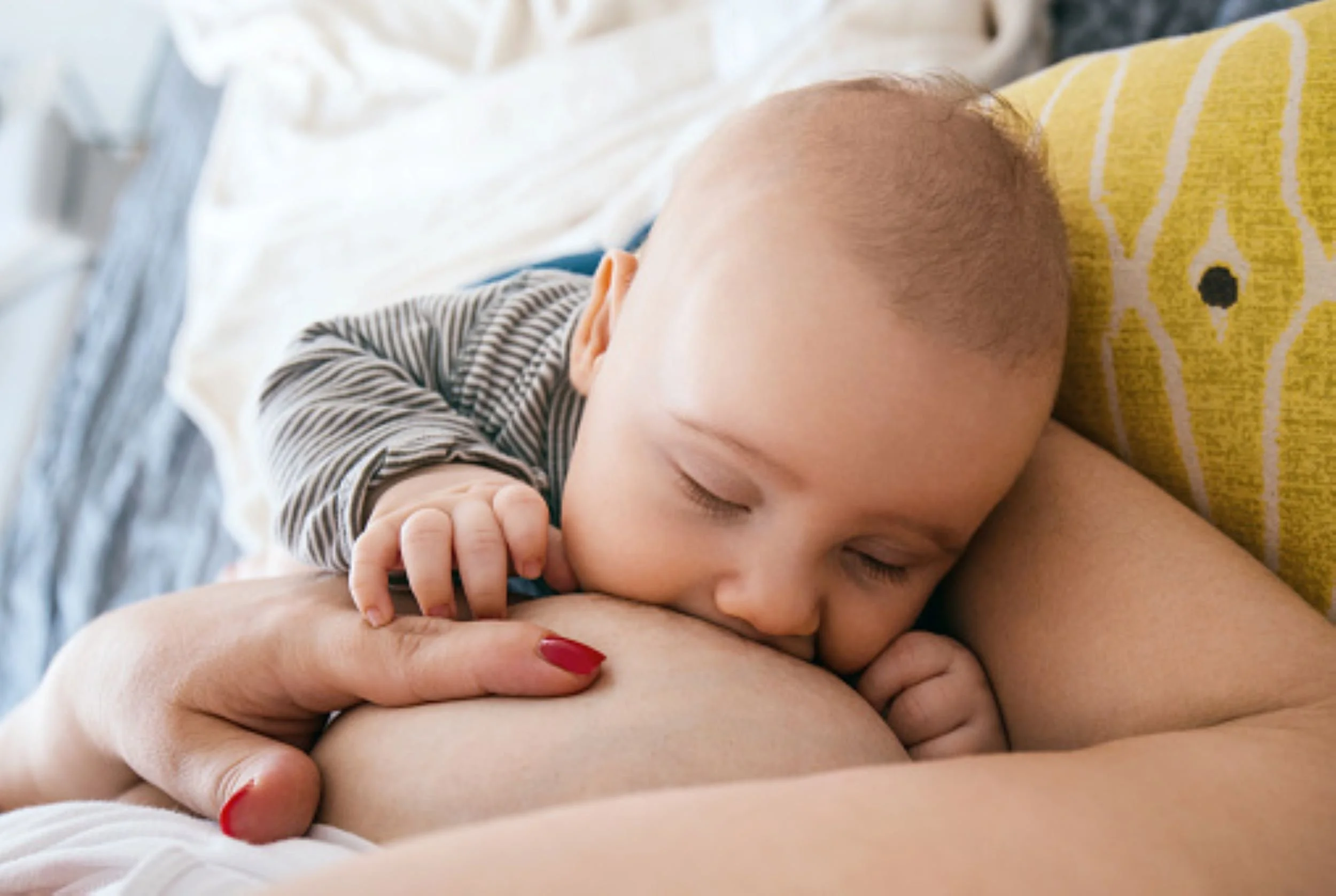 Baby lying back, latched to mother's breast, wearing a striped onesie