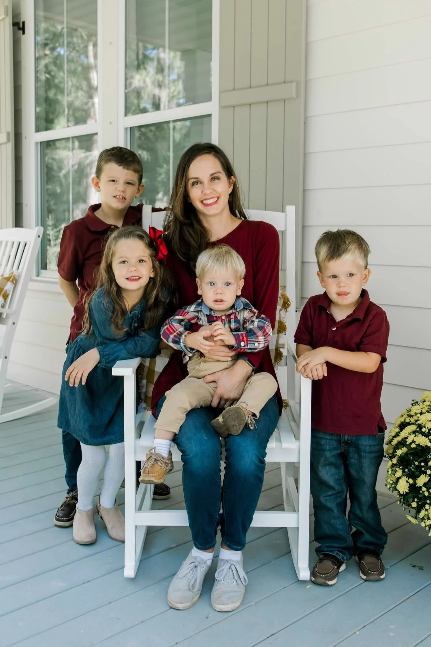 A woman with five children sitting and standing on a porch with white siding and large windows, smiling and posing for a photo.