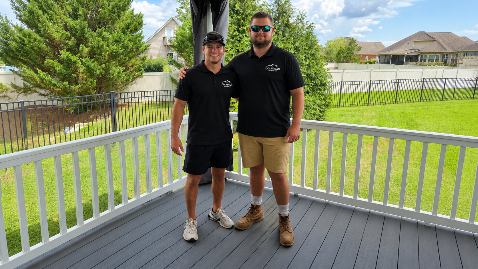 Two men standing on a porch, smiling at the camera. Both are wearing black polo shirts with a logo, one in black shorts and the other in khaki shorts and brown boots. A backyard with green grass, trees, and a white fence is visible in the background.